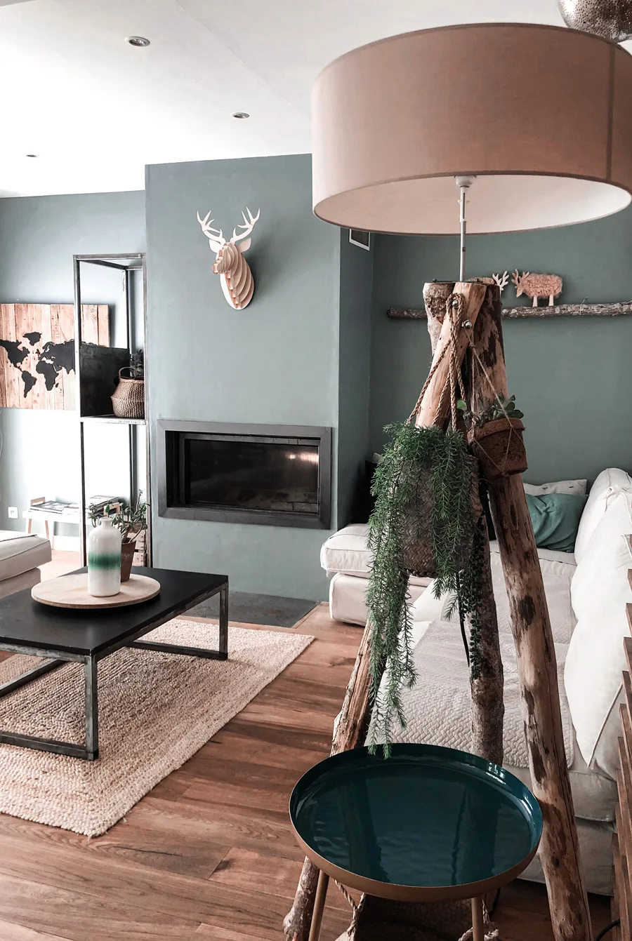 A living room featuring a sage green fireplace wall, a white sectional sofa, a black coffee table, and a unique wood log tripod lamp.