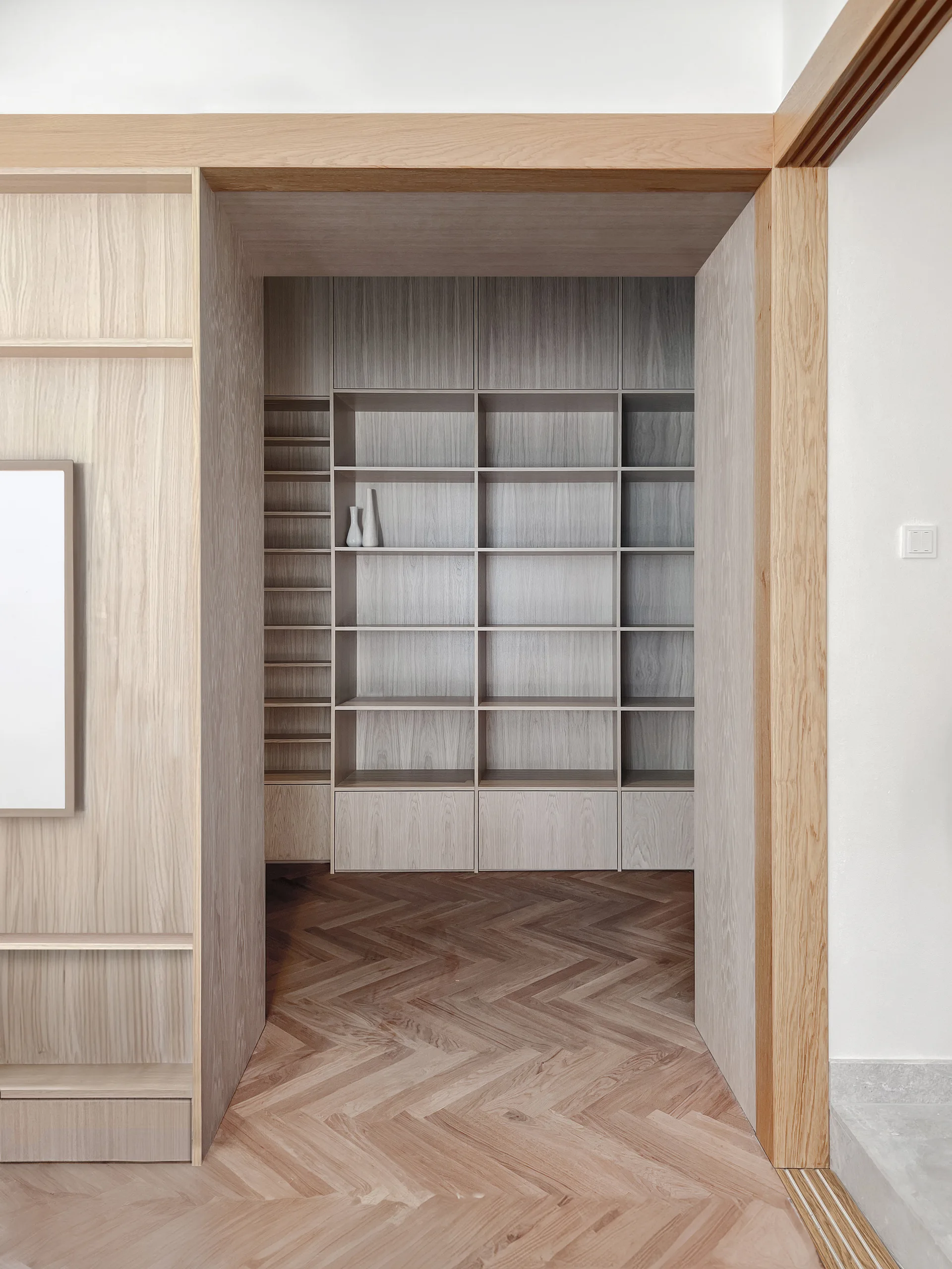 A light wood transitional entryway with a wide timber-framed threshold opening onto a minimalist built-in bookcase and herringbone pattern flooring.