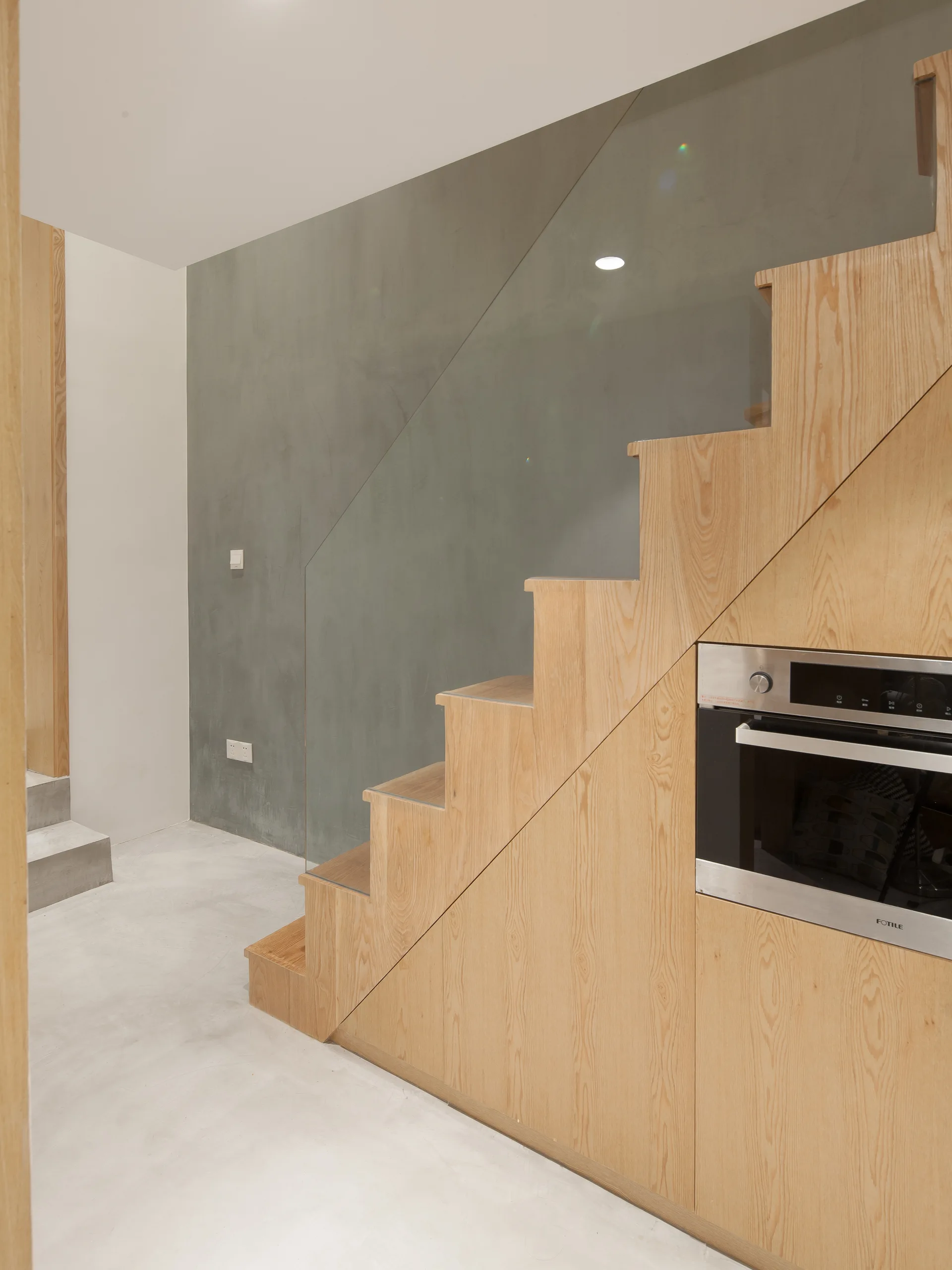 A light wood staircase features a clear glass railing and a stainless steel oven integrated into the cabinetry under the stairs.