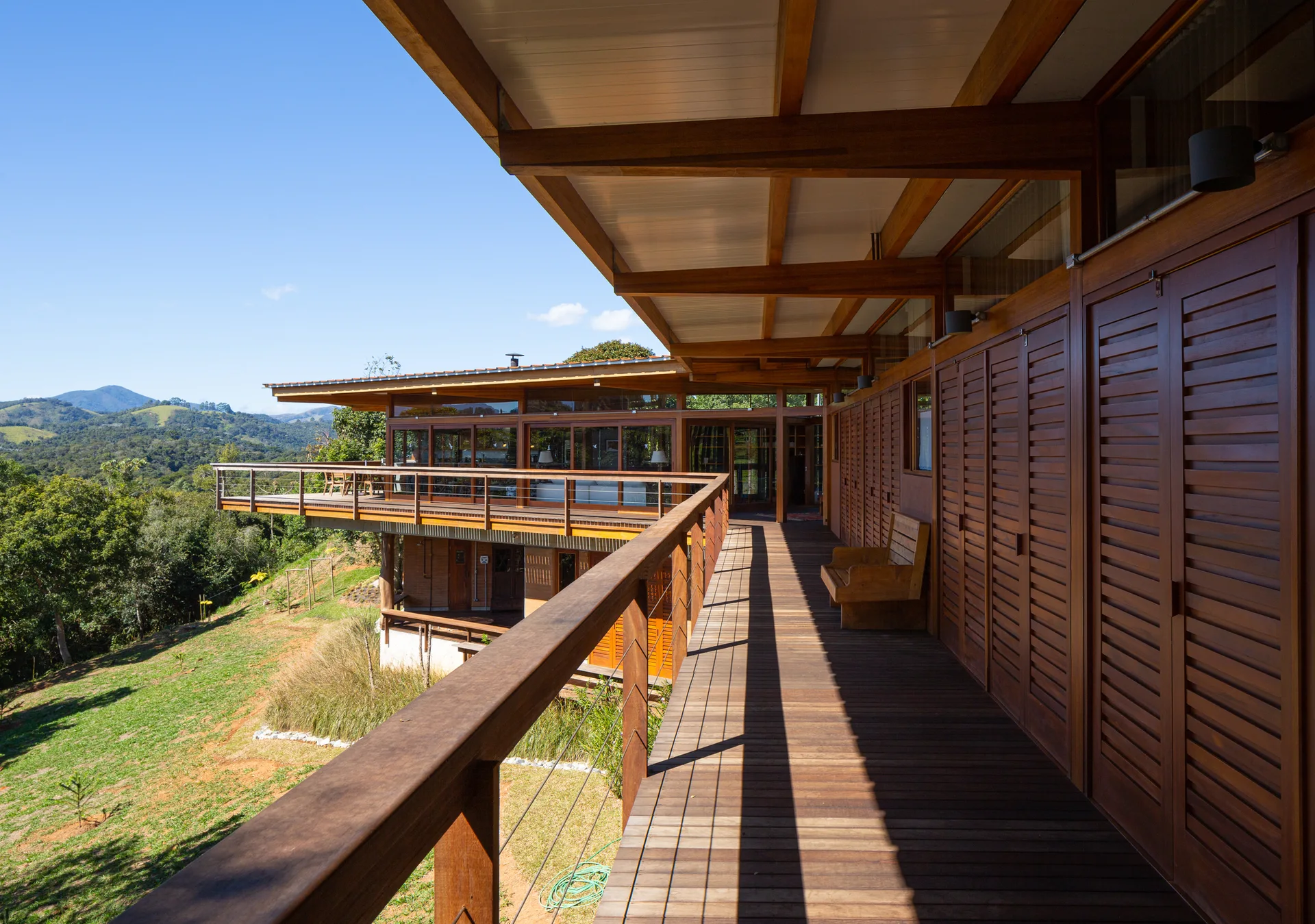 A long covered wooden balcony with exposed ceiling beams and a wall of rich brown louvered shutters overlooks a green hilly landscape.