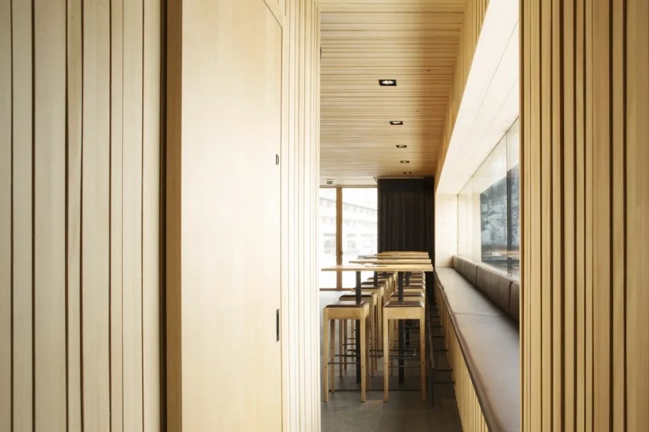 A long narrow seating area with vertical wood slat paneling on the walls and ceiling, featuring a continuous banquette and high bar stools.