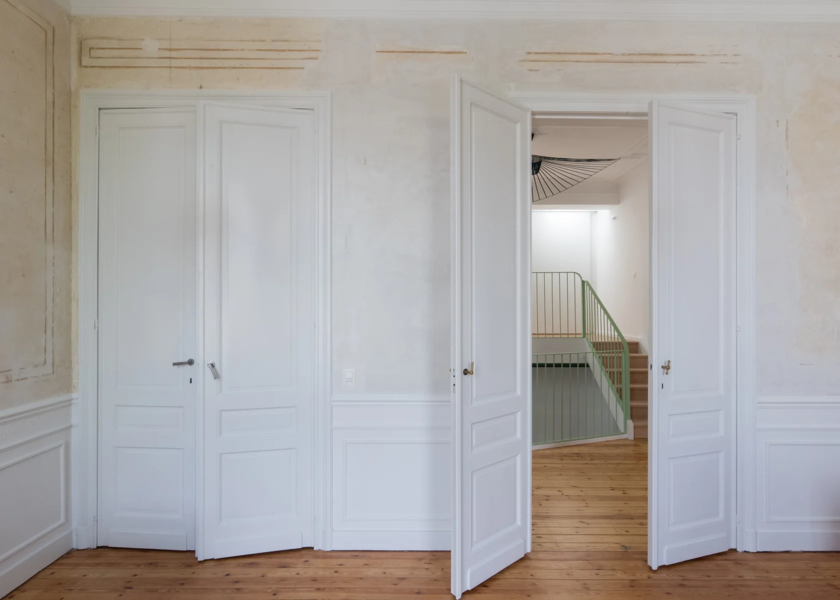 White paneled double doors open to reveal a modern staircase with a light green metal railing, set against distressed plaster walls and wood floors.