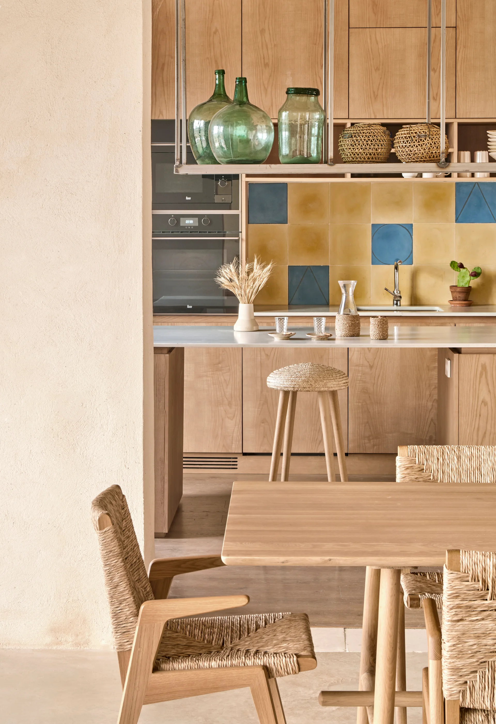 A light wood kitchen features a yellow and blue tile backsplash, a central island with a woven stool, and wooden dining chairs in the foreground.