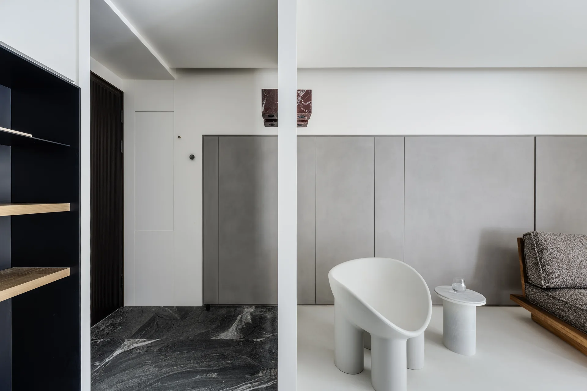 A minimalist living space featuring a white sculptural armchair, a small marble side table, grey wall paneling, and a split dark stone floor.
