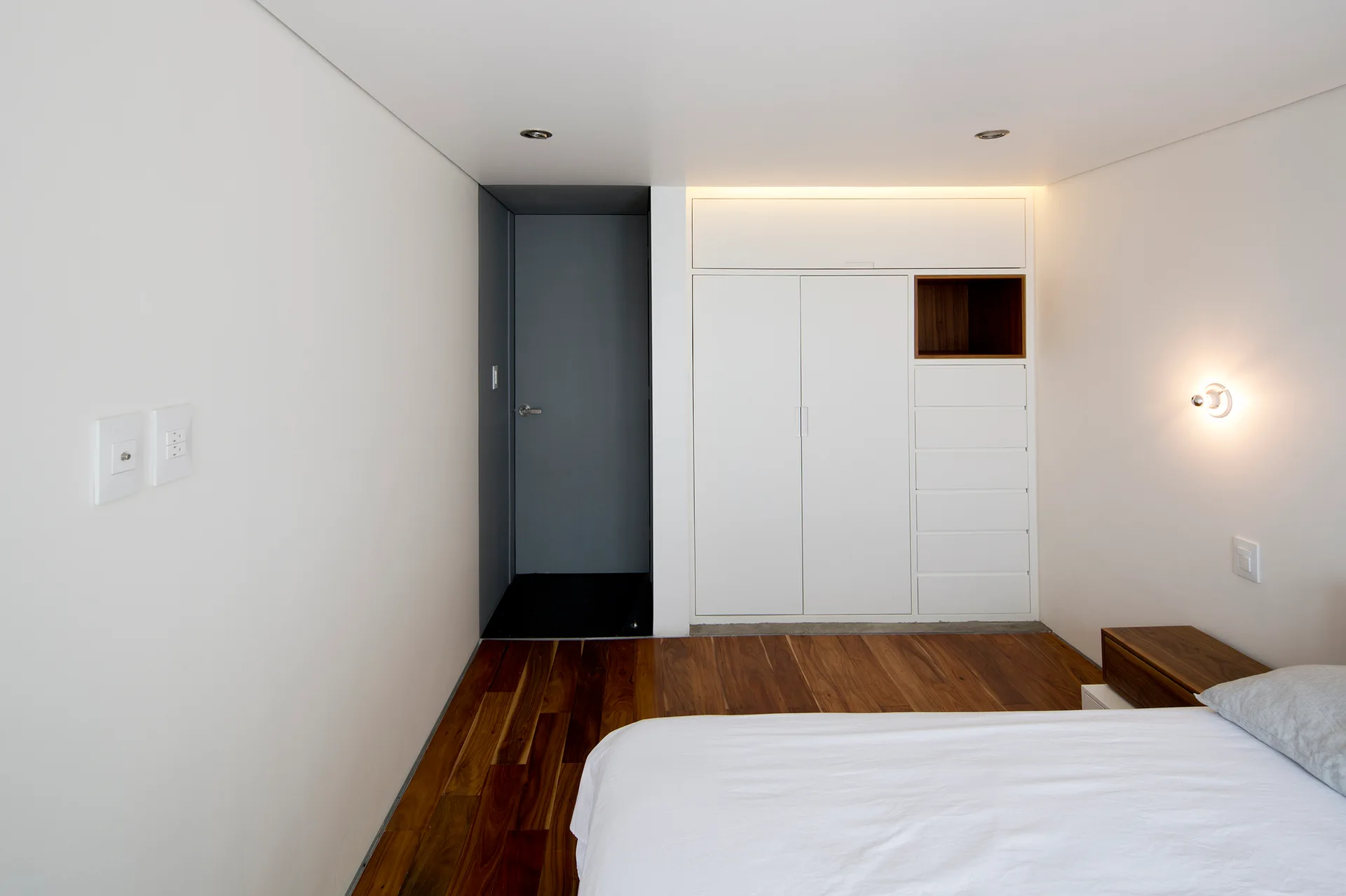 A minimalist bedroom featuring white walls, a dark grey doorway, a built-in white wardrobe with a wood cubby, and warm hardwood floors.