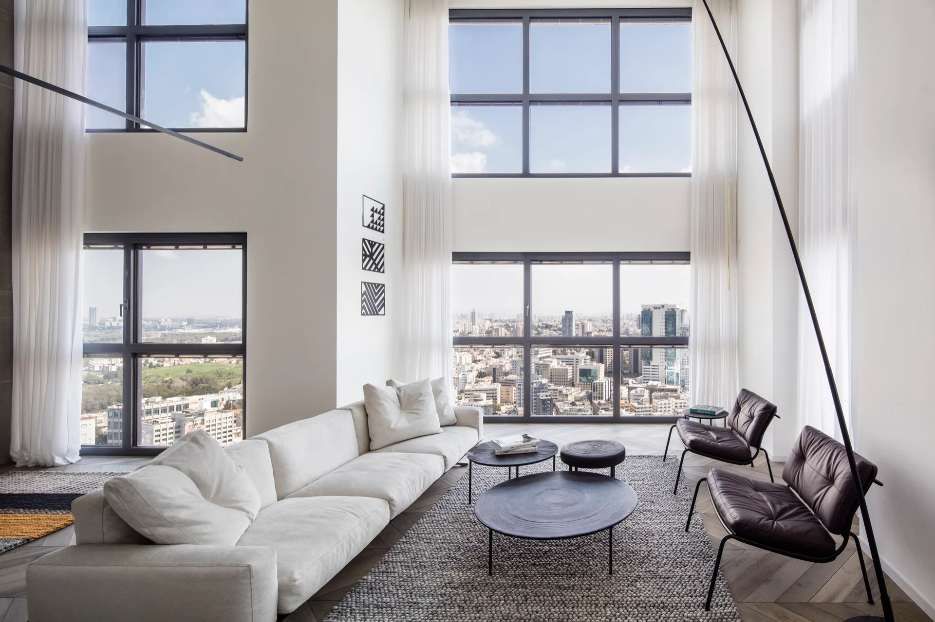 A high-ceilinged modern living room features a long white modular sofa, two dark leather armchairs, and floor-to-ceiling windows showing a city skyline.