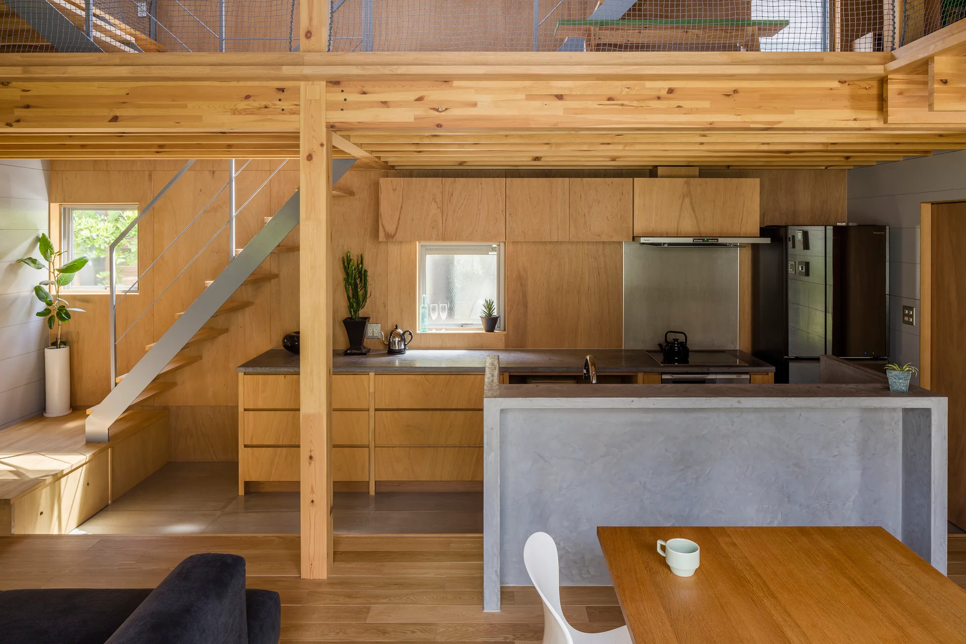 A modern minimalist kitchen with light wood paneled walls, a gray concrete kitchen island, and an open staircase leading to a mezzanine level.