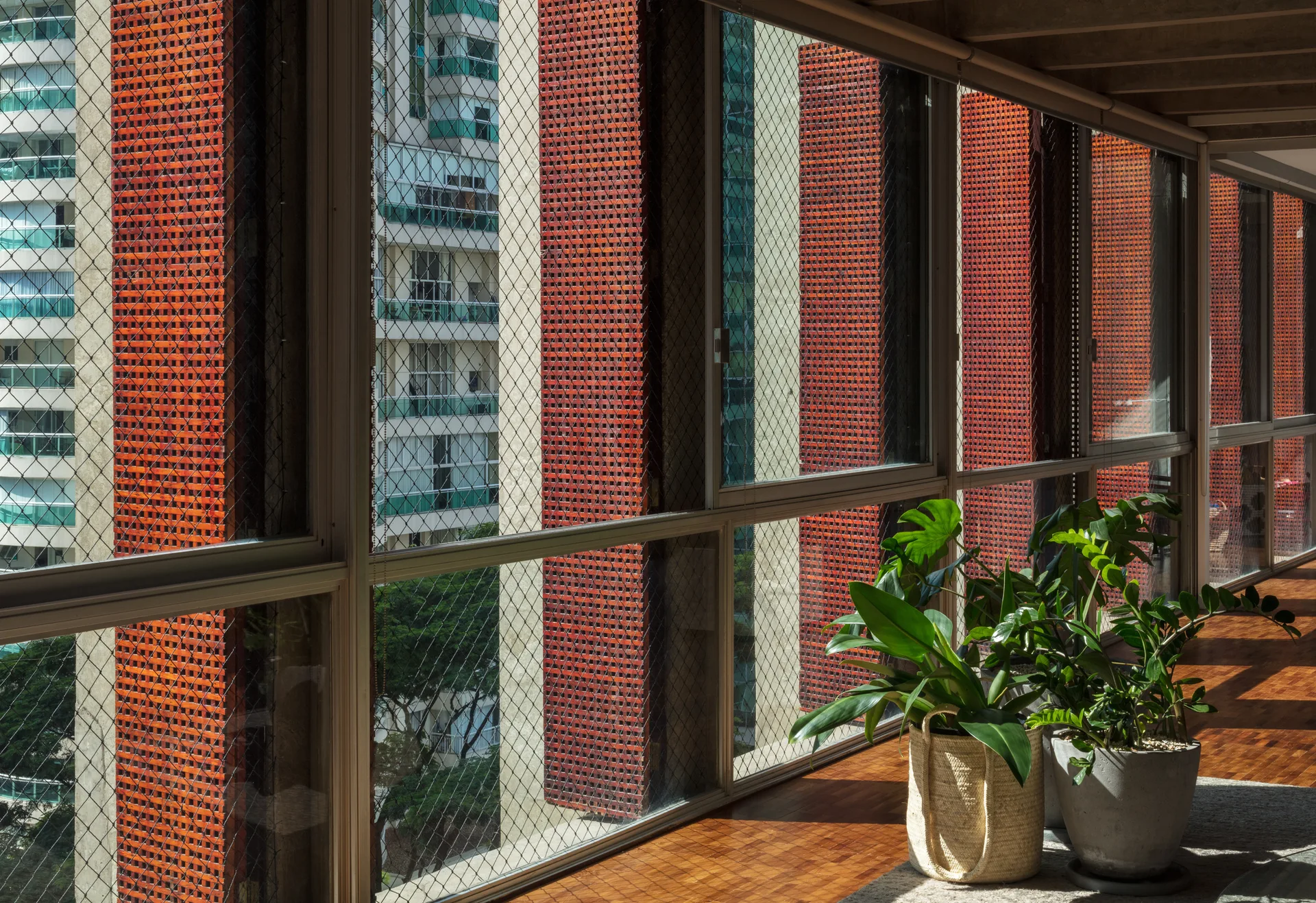 An enclosed balcony features parquet flooring, large windows overlooking a city, and green potted plants in a woven basket and concrete planter.