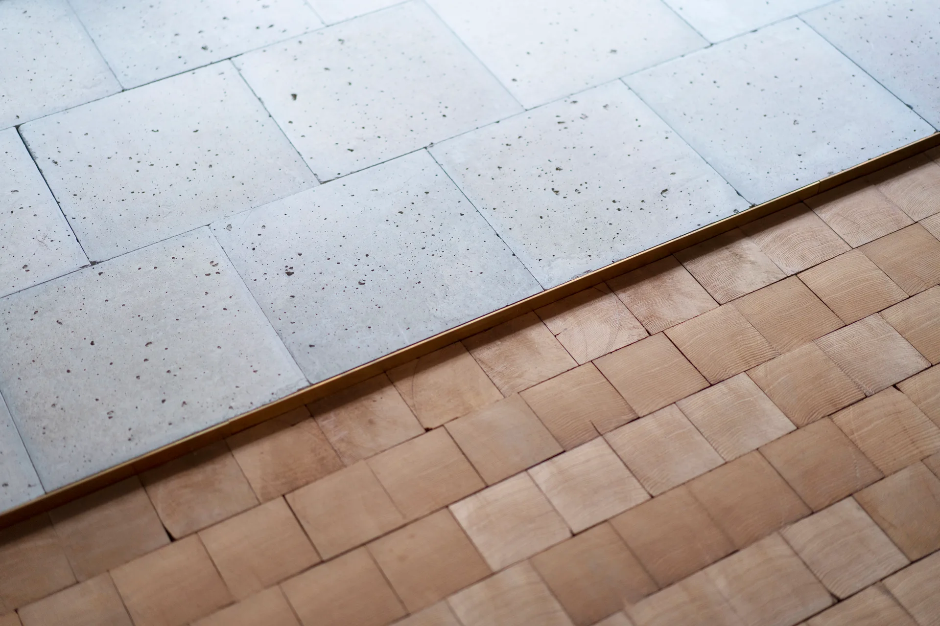 A close-up of a floor transition between pitted gray concrete tiles and tan wood end-grain blocks, divided by a thin brass metal strip.