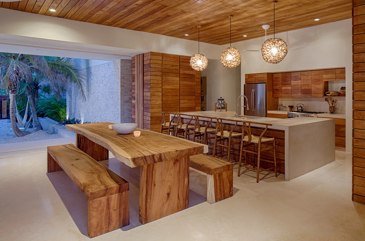 A modern open-plan kitchen featuring a wood ceiling, concrete island, wooden bar stools, and a large live-edge wood dining table next to an outdoor patio.