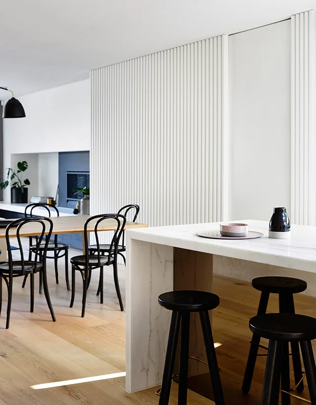 A marble waterfall kitchen island with black stools sits in front of a dining table with bentwood chairs and a white fluted wall.