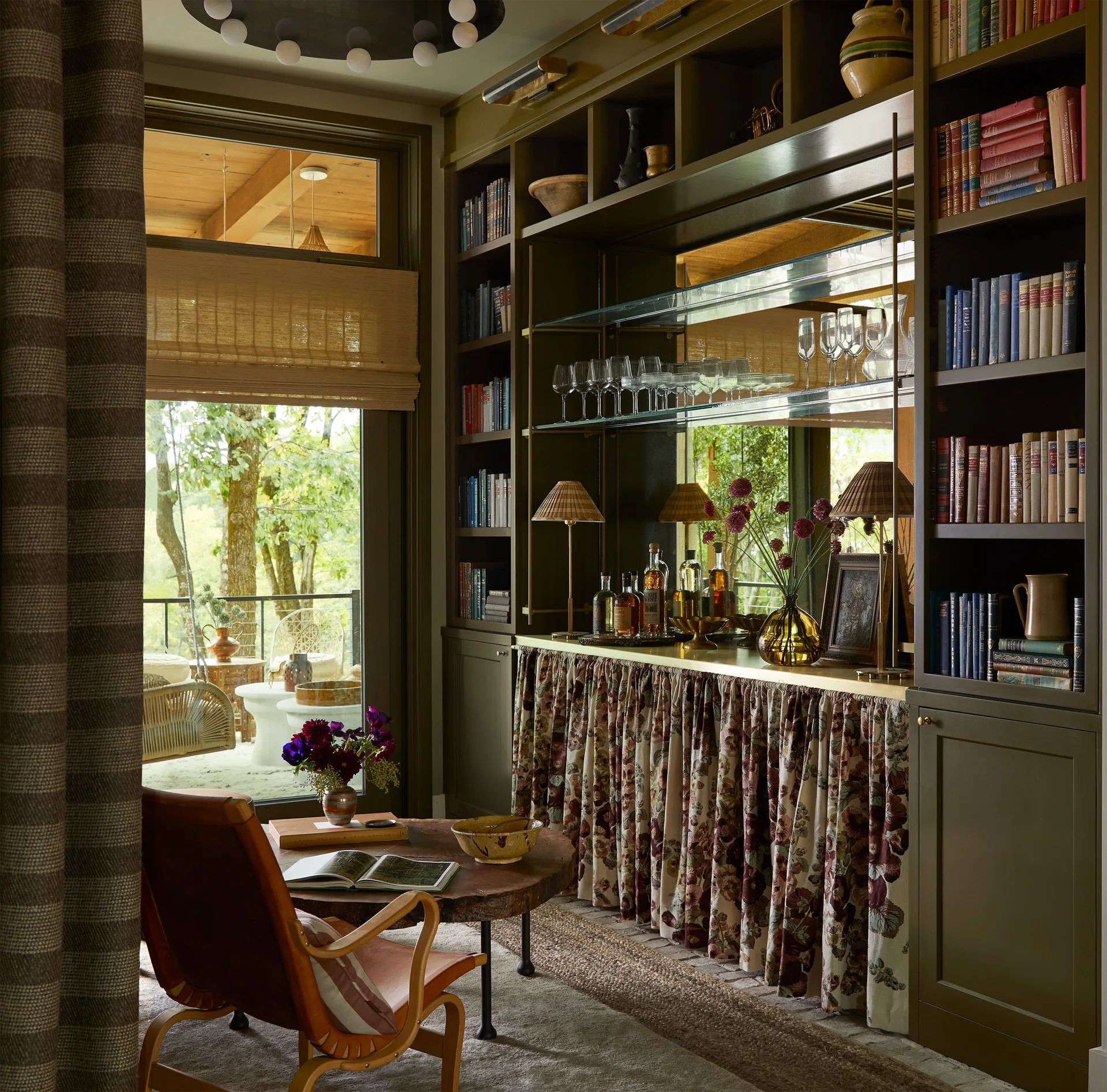 Olive green built-in bookshelves feature an integrated mirrored bar with glassware and a floral skirt beside a leather armchair and live-edge table.