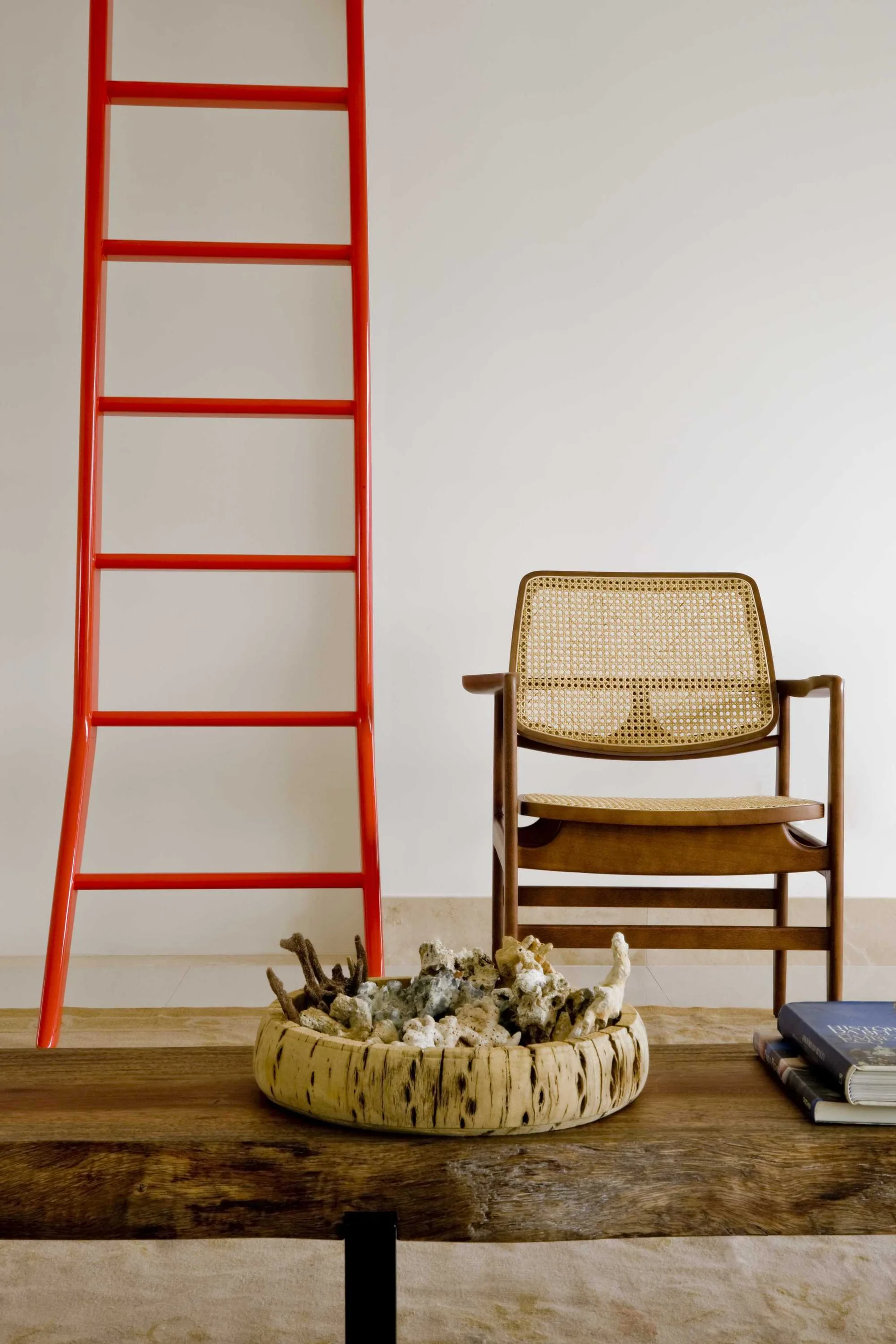 A minimalist living room vignette featuring a wooden armchair with a woven cane back next to a bright red leaning ladder.