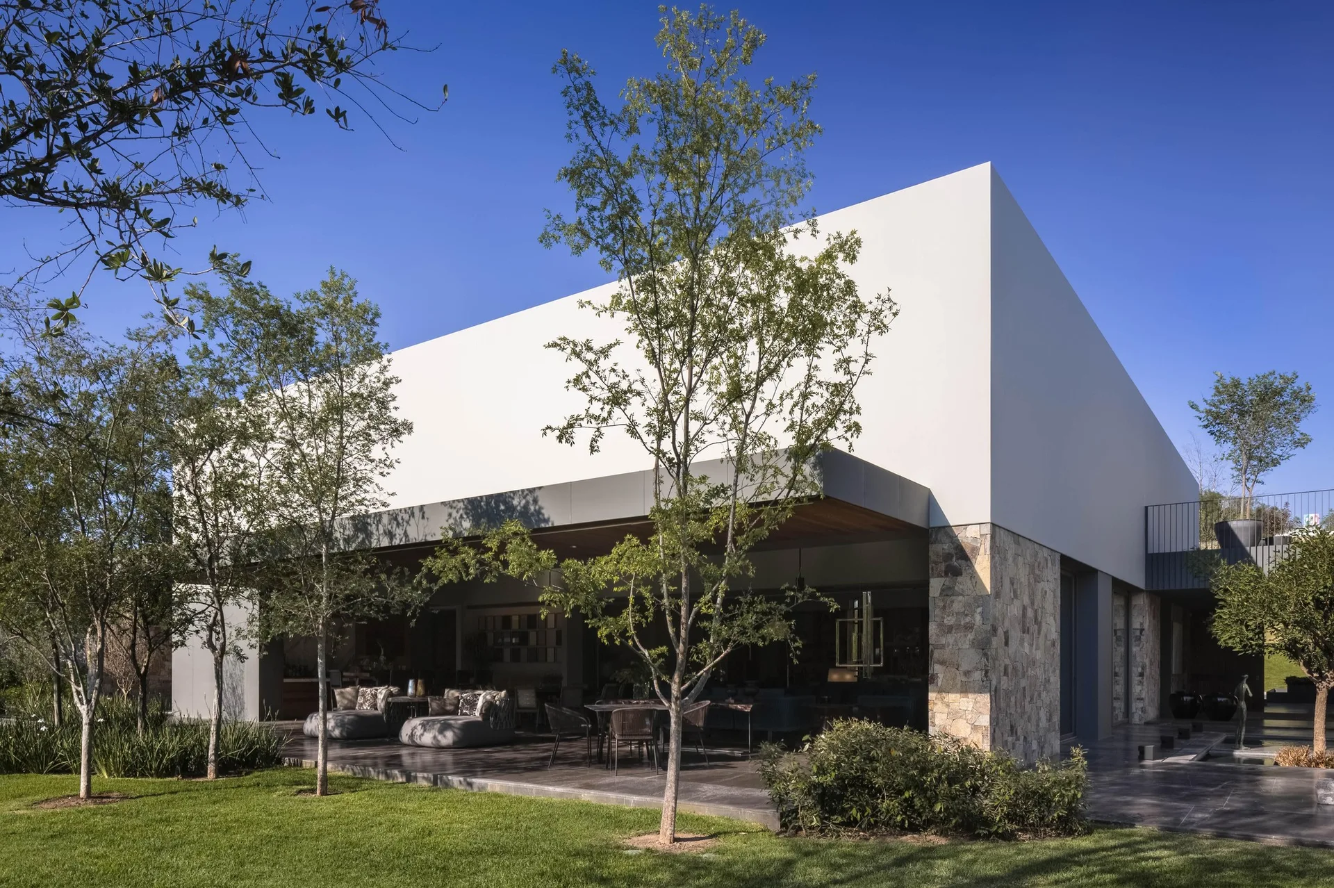 A modern home exterior features a deep covered patio with gray outdoor furniture, a wood plank ceiling, a stone masonry wall, and a green lawn.
