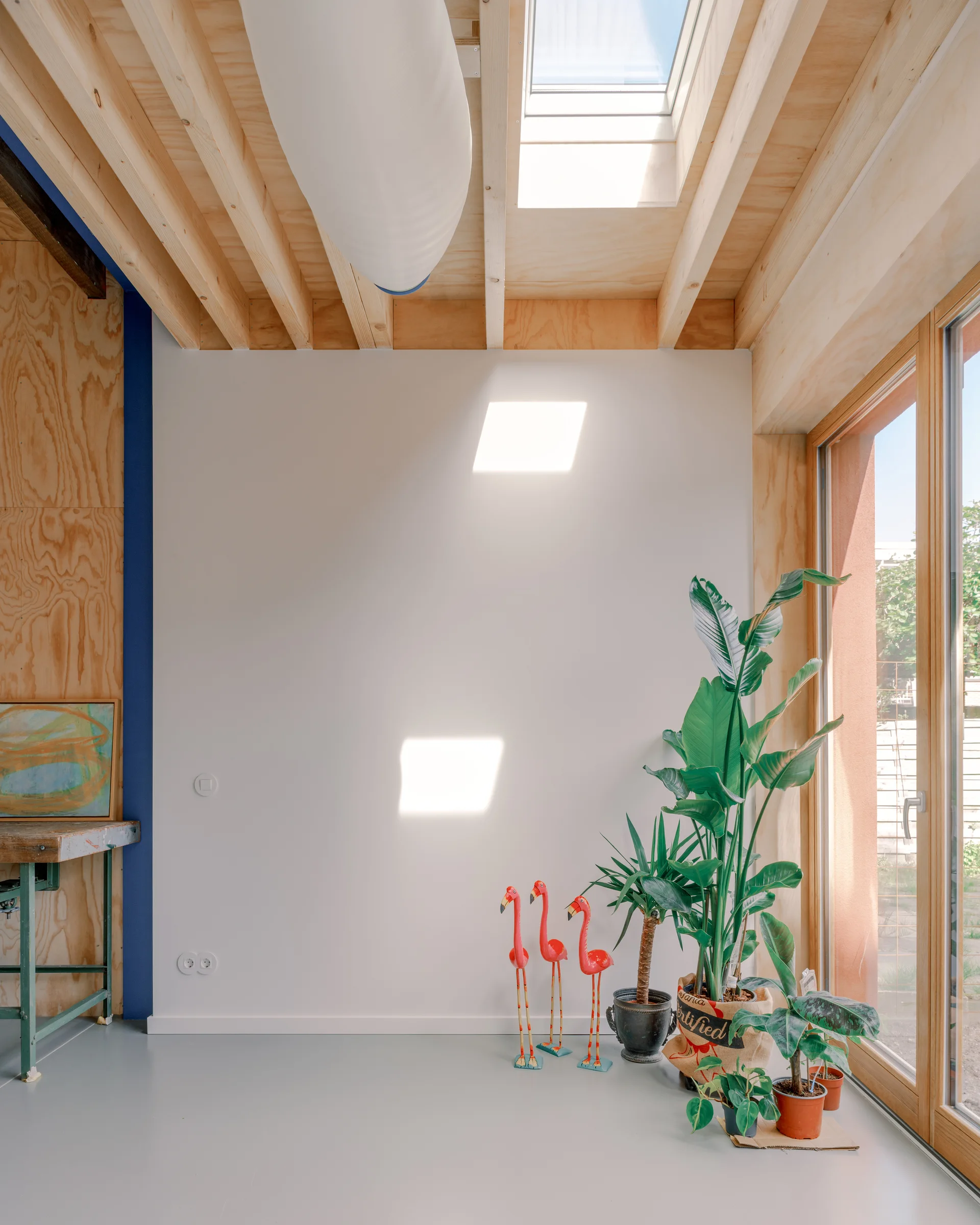 A sunlit room featuring exposed plywood ceiling joists, a skylight, a large glass door, and potted plants clustered next to three flamingo figurines.