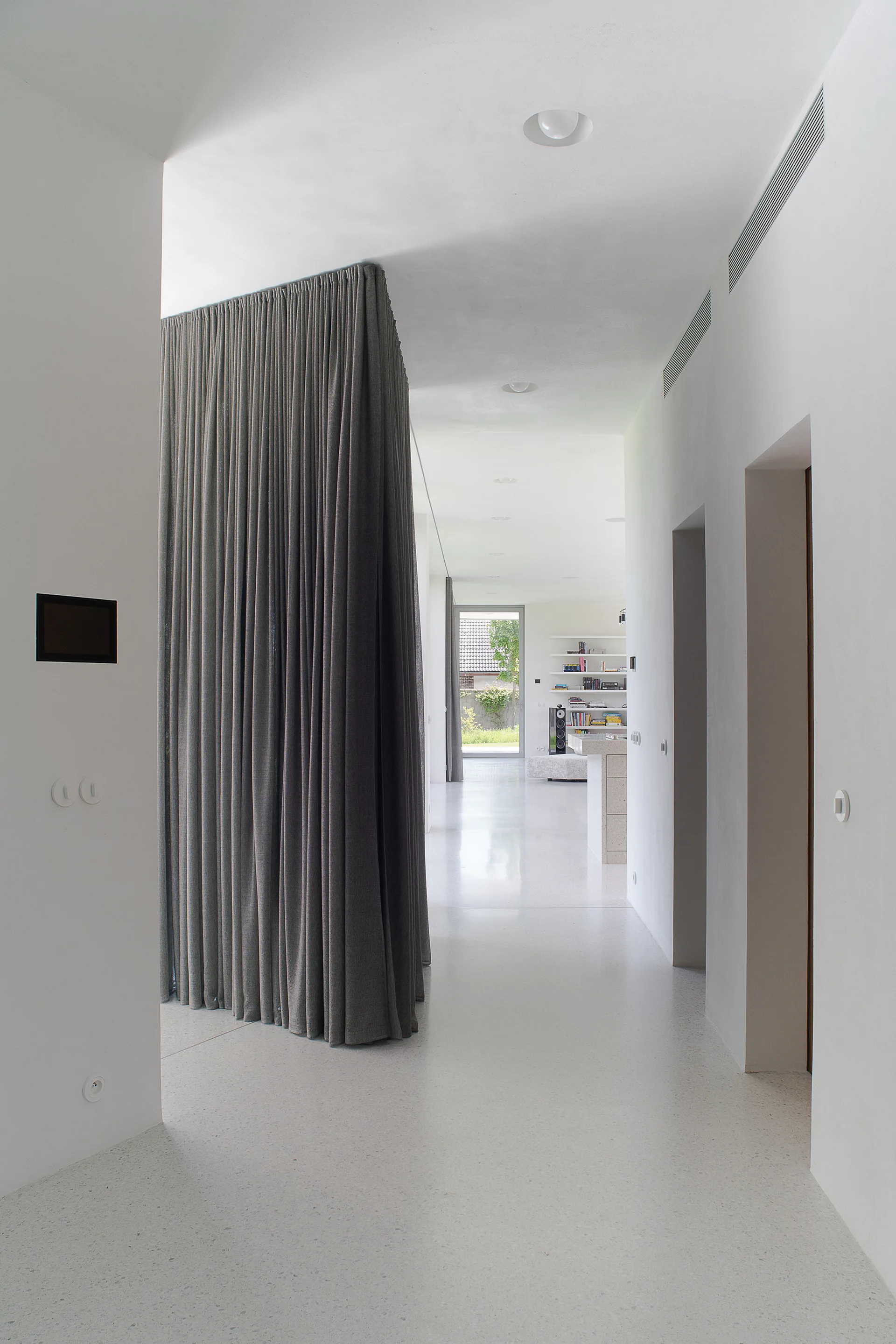 A minimalist white hallway featuring a large floor-to-ceiling grey fabric curtain partition and smooth white terrazzo floors.