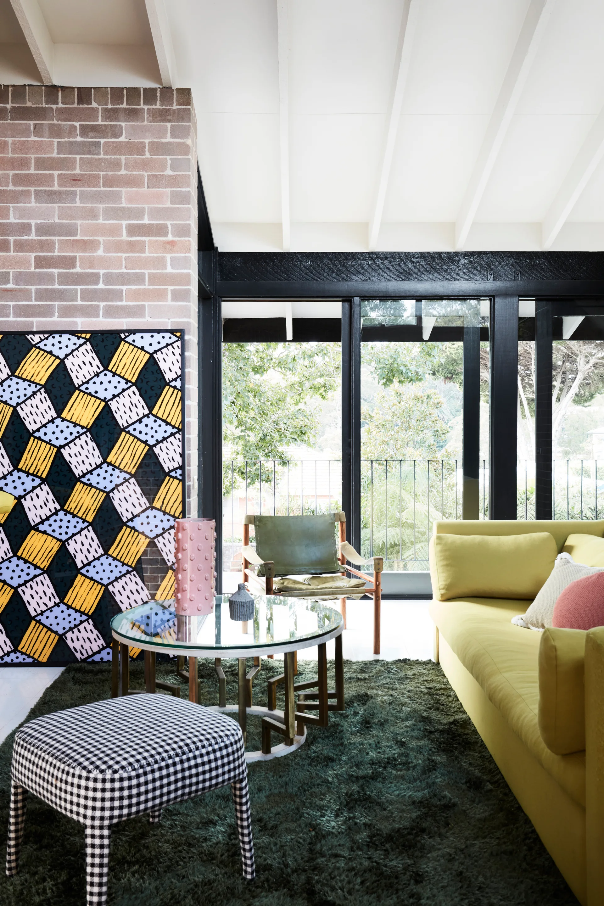 An eclectic living room features a yellow sofa, a checkered ottoman, and a glass coffee table in front of a brick fireplace.