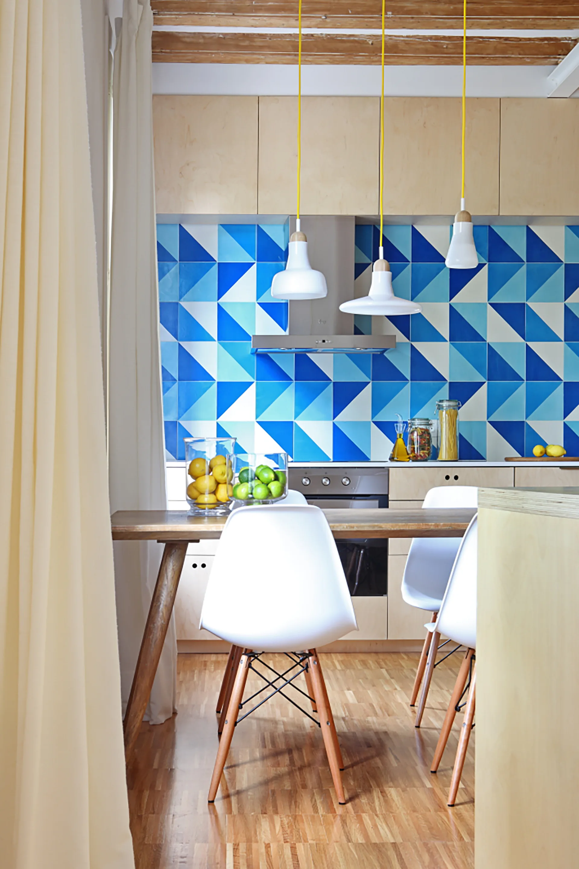A Scandinavian-style kitchen features a bold blue geometric tile backsplash, light wood cabinetry, and a dining table with white chairs.