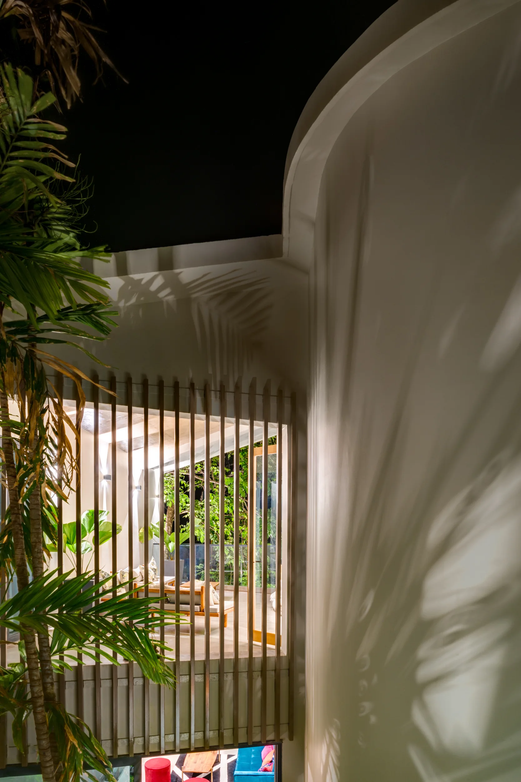 A night view of a tropical terrace behind a wooden slatted screen, featuring a curved white wall with cast palm leaf shadows and outdoor seating.