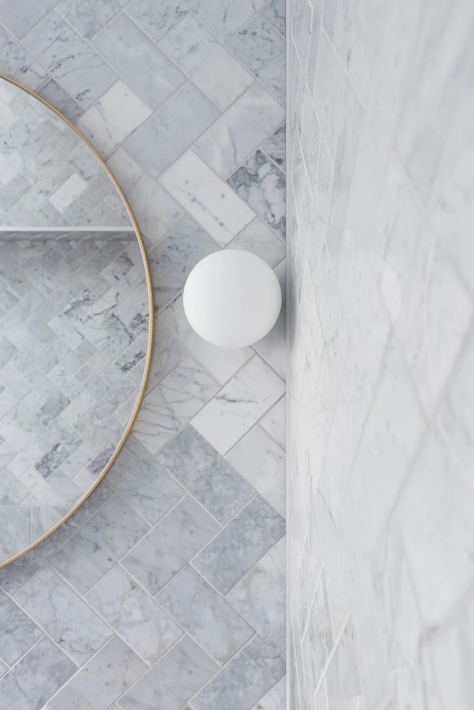 A close-up of a bathroom wall features white marble herringbone tiles, a thin brass-rimmed circular mirror, and a white spherical sconce.