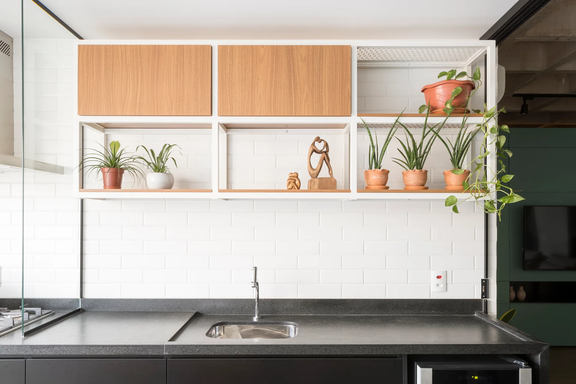 A dark stone kitchen countertop with a metal sink sits beneath white subway tile and custom white metal open shelving displaying potted plants.