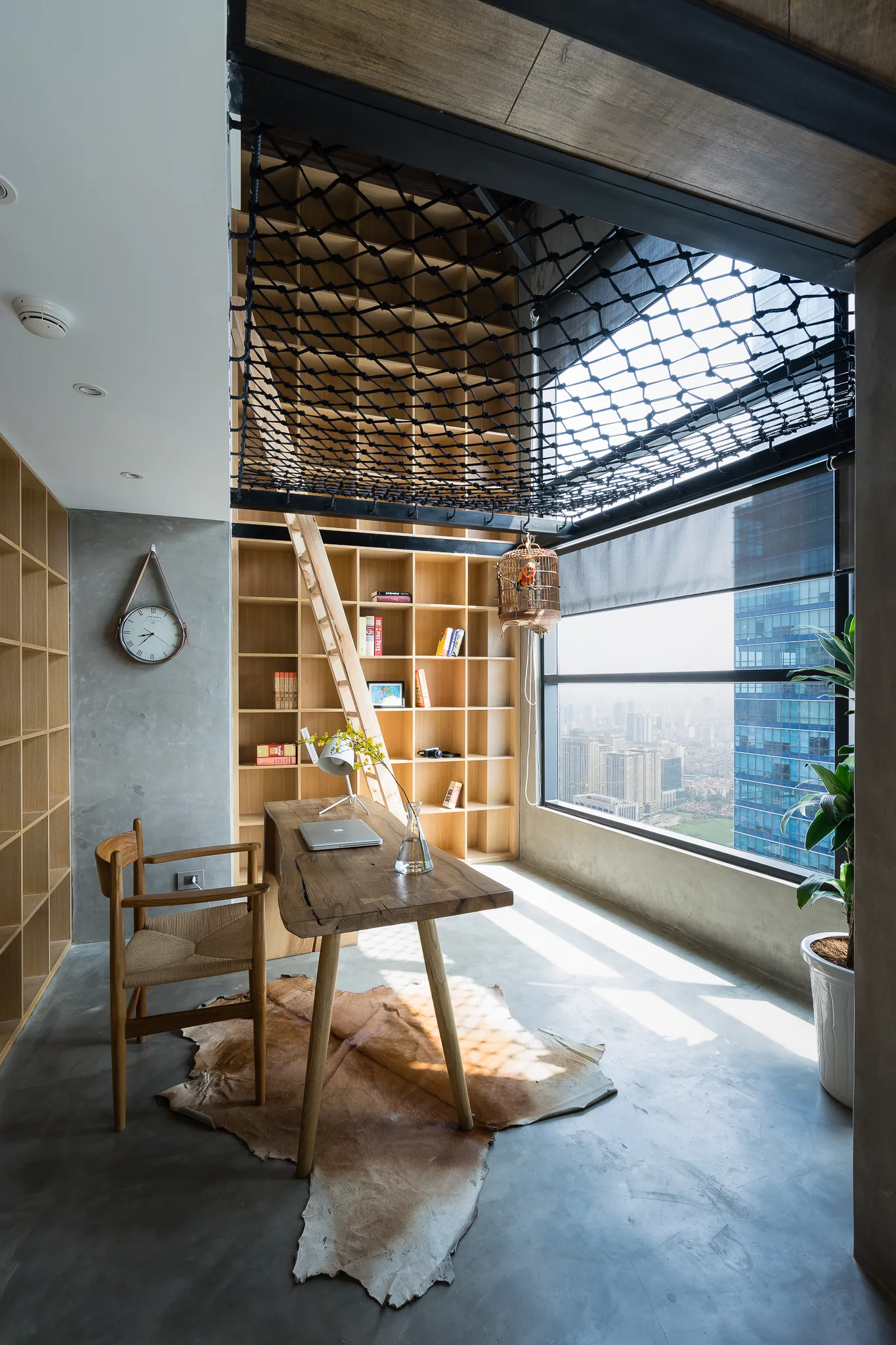 A modern home office featuring concrete walls, a natural wood desk, built-in floor-to-ceiling bookshelves with a ladder, and a black cargo net loft overhead.