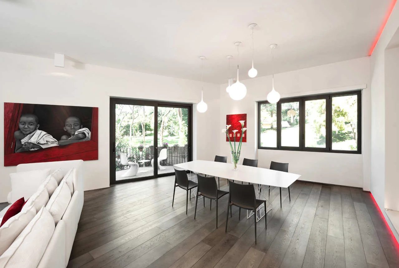 A minimalist dining room featuring a long white table, black chairs, a cluster of globe pendant lights, and red accent artwork.