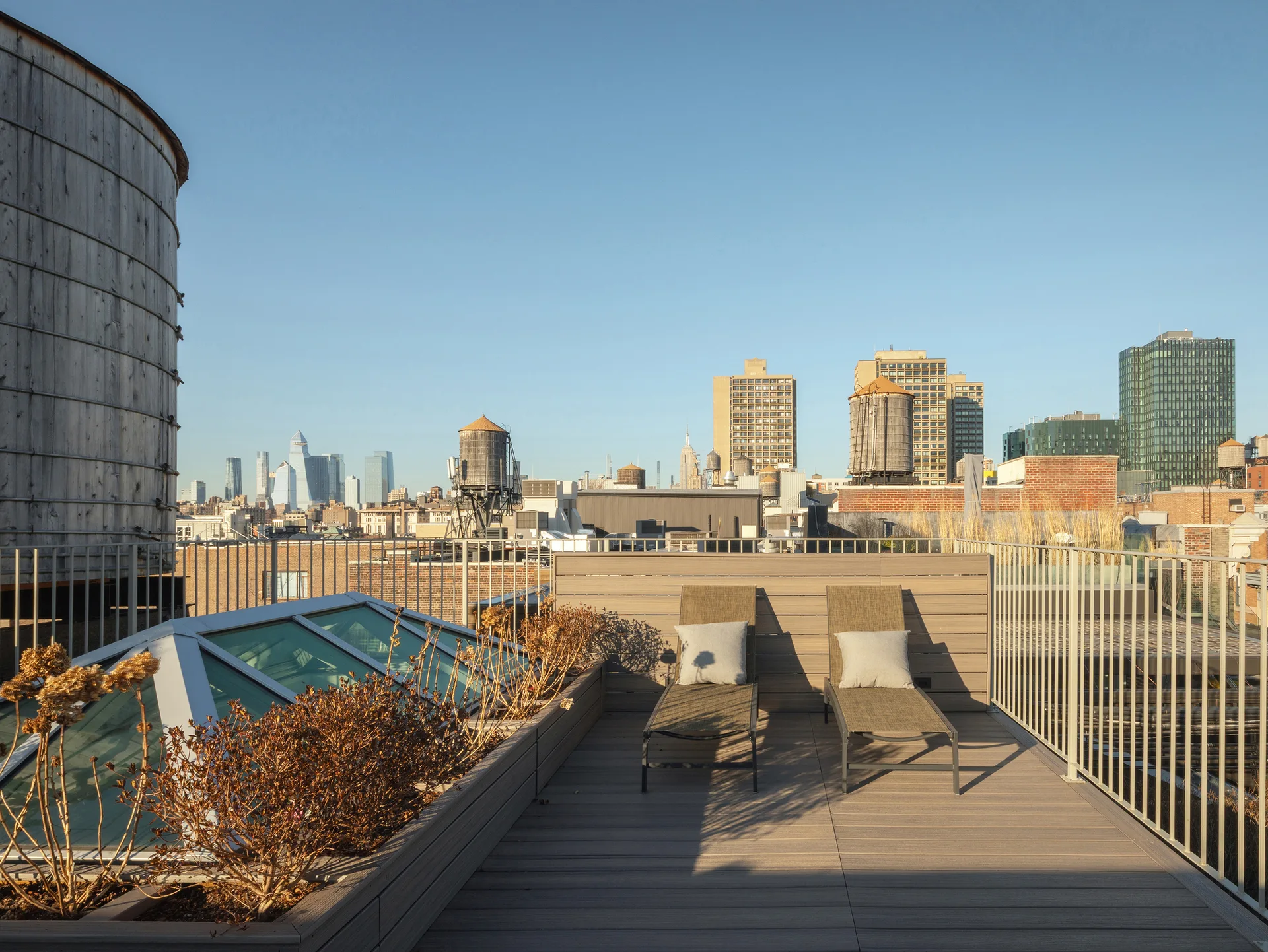 An urban rooftop terrace features two brown lounge chairs on a wood-look deck next to a glass skylight with city buildings in the background.