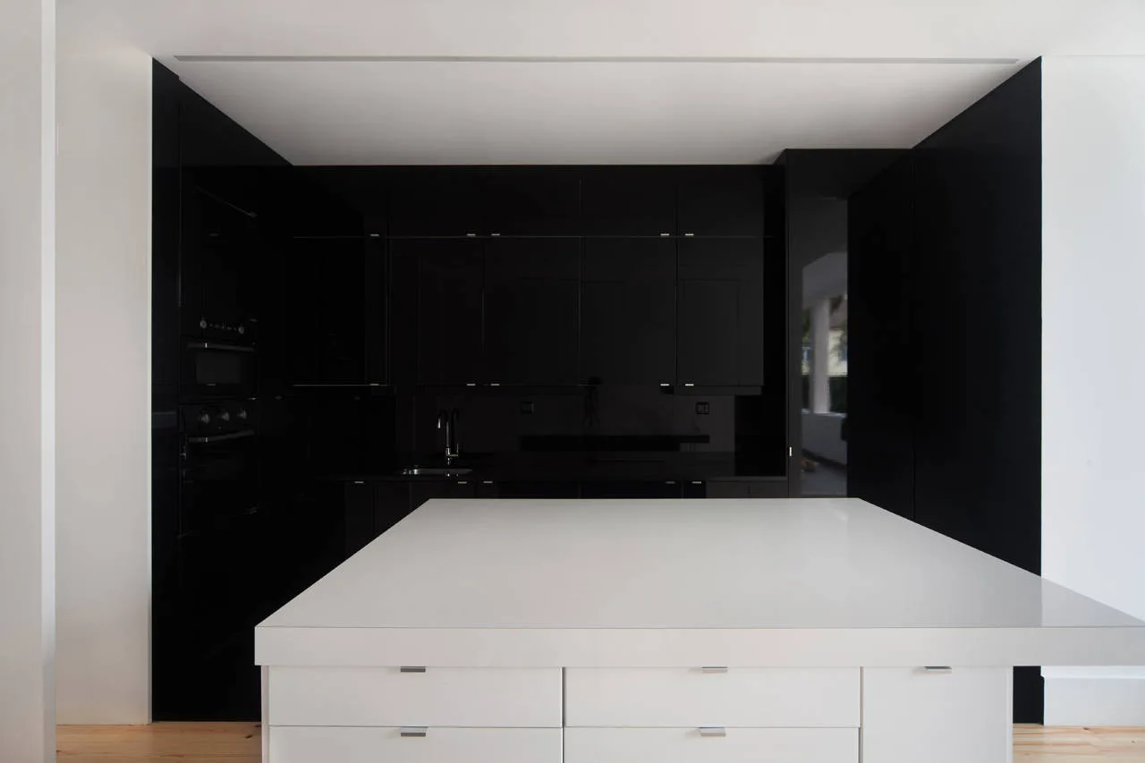 A minimalist kitchen features a large white central island with drawers in front of seamless glossy black back cabinetry.