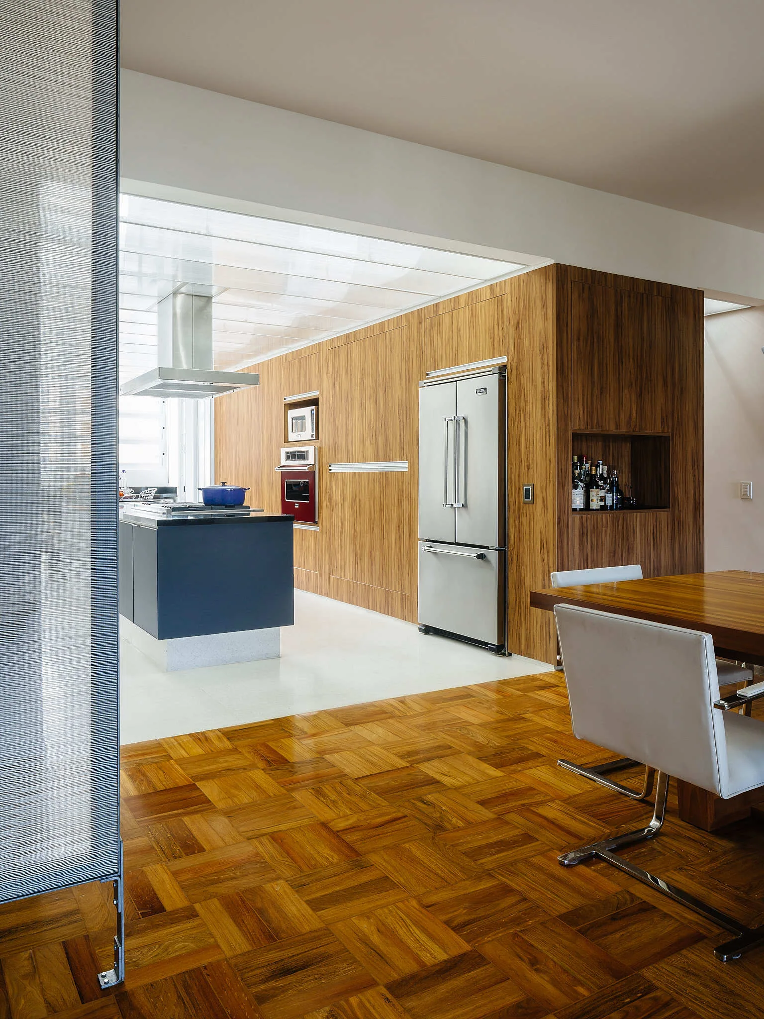A modern kitchen with a blue island, wood-paneled wall cabinetry, integrated stainless steel refrigerator, and parquet flooring in the dining area.