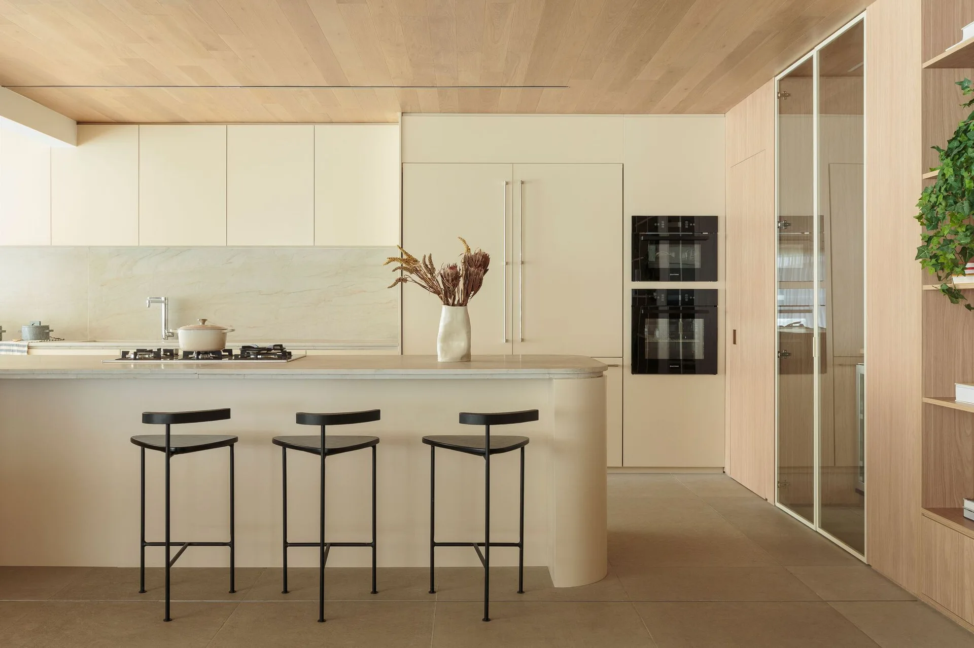 A minimalist kitchen features a curved beige island with three black stools, light wood ceiling planks, and pale integrated cabinets.