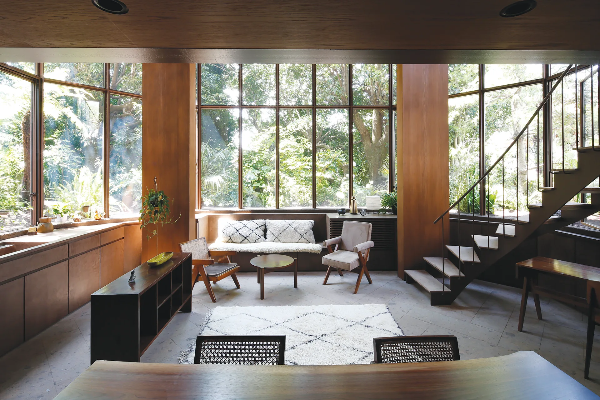 A wood-paneled living room features mid-century modern armchairs, an open-tread staircase, and large grid windows overlooking a lush garden.