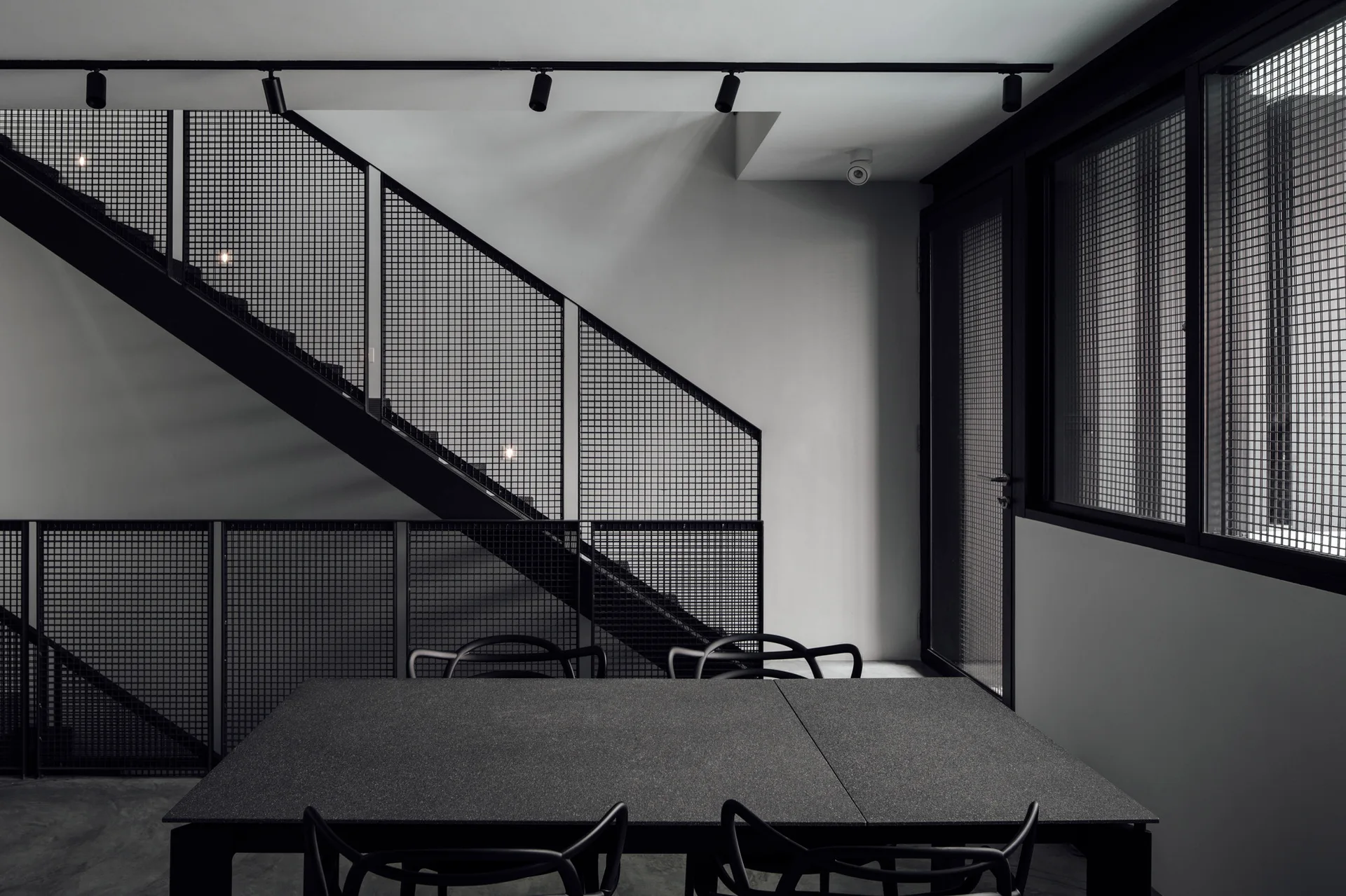 A minimalist dining room features a dark gray table with black curved chairs below a steel staircase with black metal mesh railings.