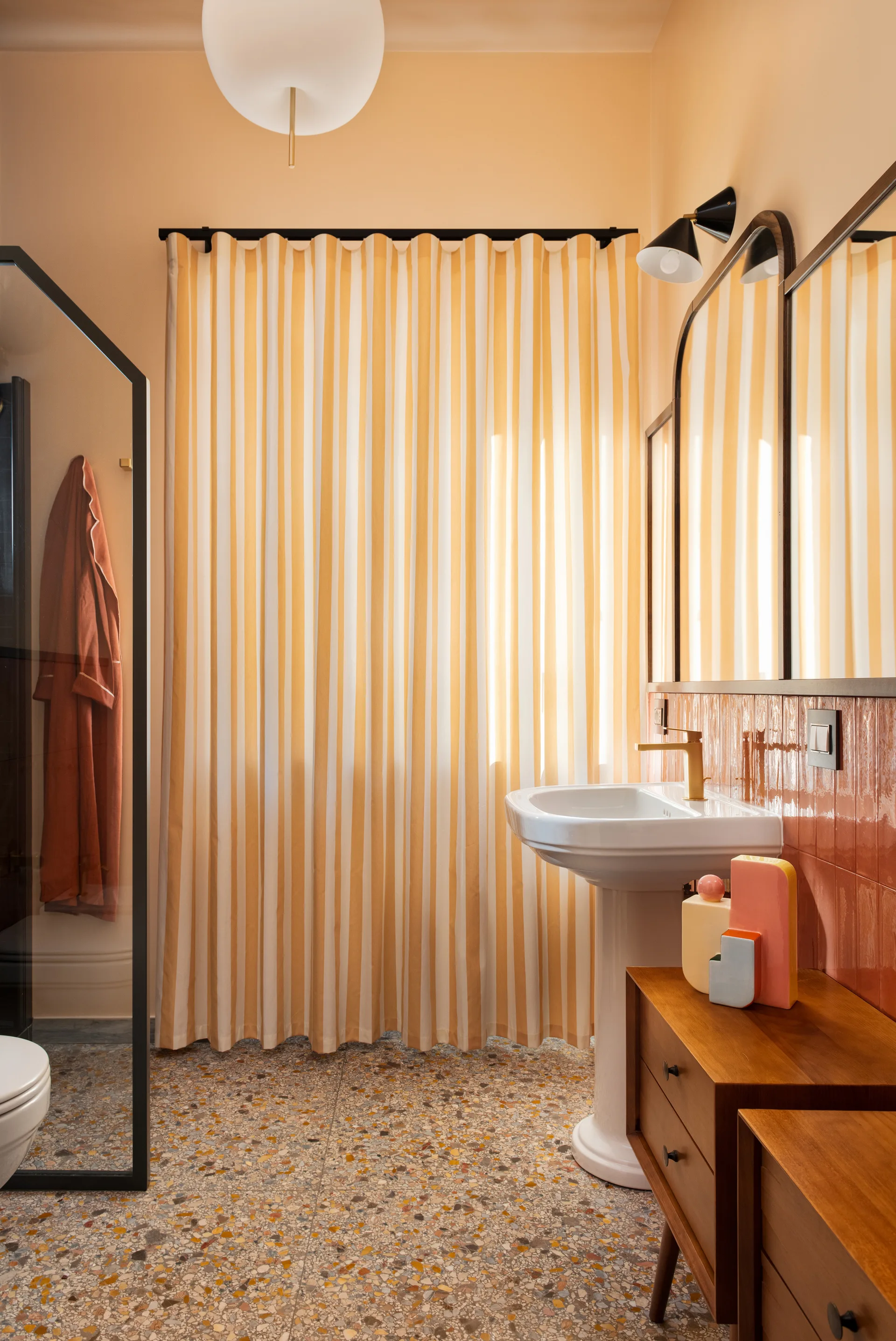 A bathroom featuring a white pedestal sink, a yellow and white striped curtain, terracotta wall tiles, and speckled terrazzo flooring.