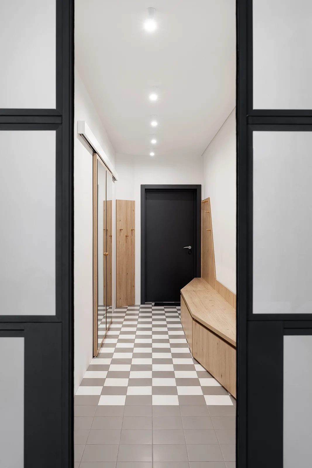 A minimalist entryway features a grey and white checkerboard tiled floor, a light wood storage bench, and a black accent door.