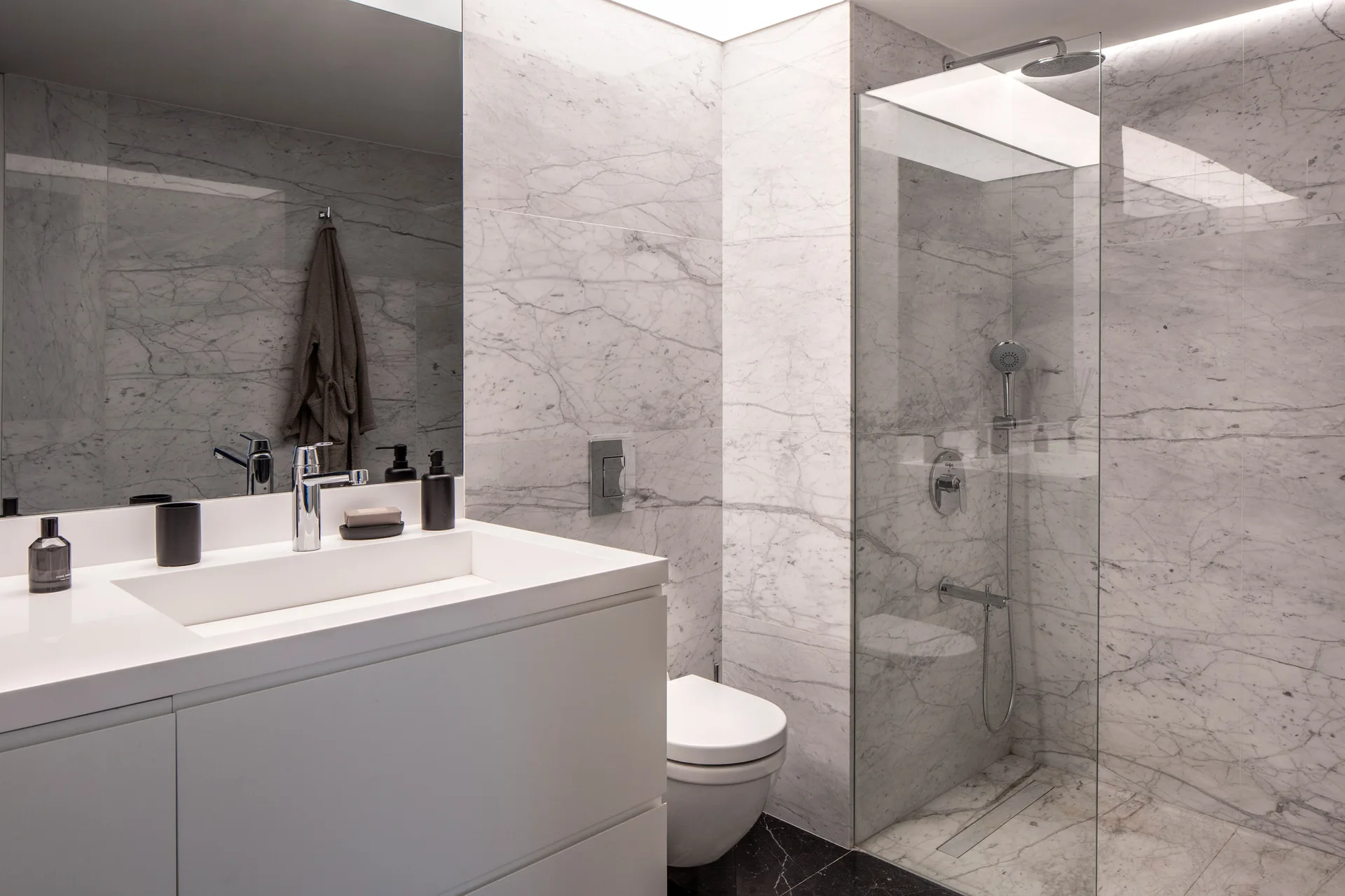 A contemporary bathroom features veined marble walls, a white floating vanity with a large mirror, a wall-hung toilet, and a glass shower enclosure.
