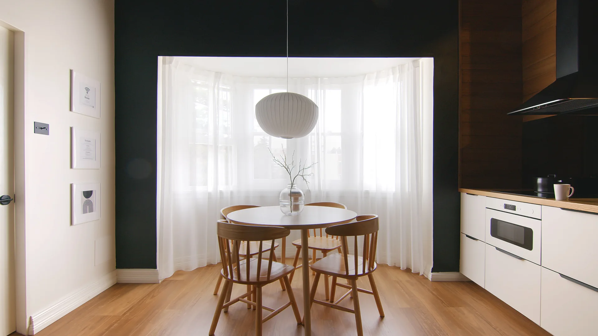 A circular wooden dining table and matching chairs are centered in front of a bay window with sheer curtains, framed by a black accent wall.