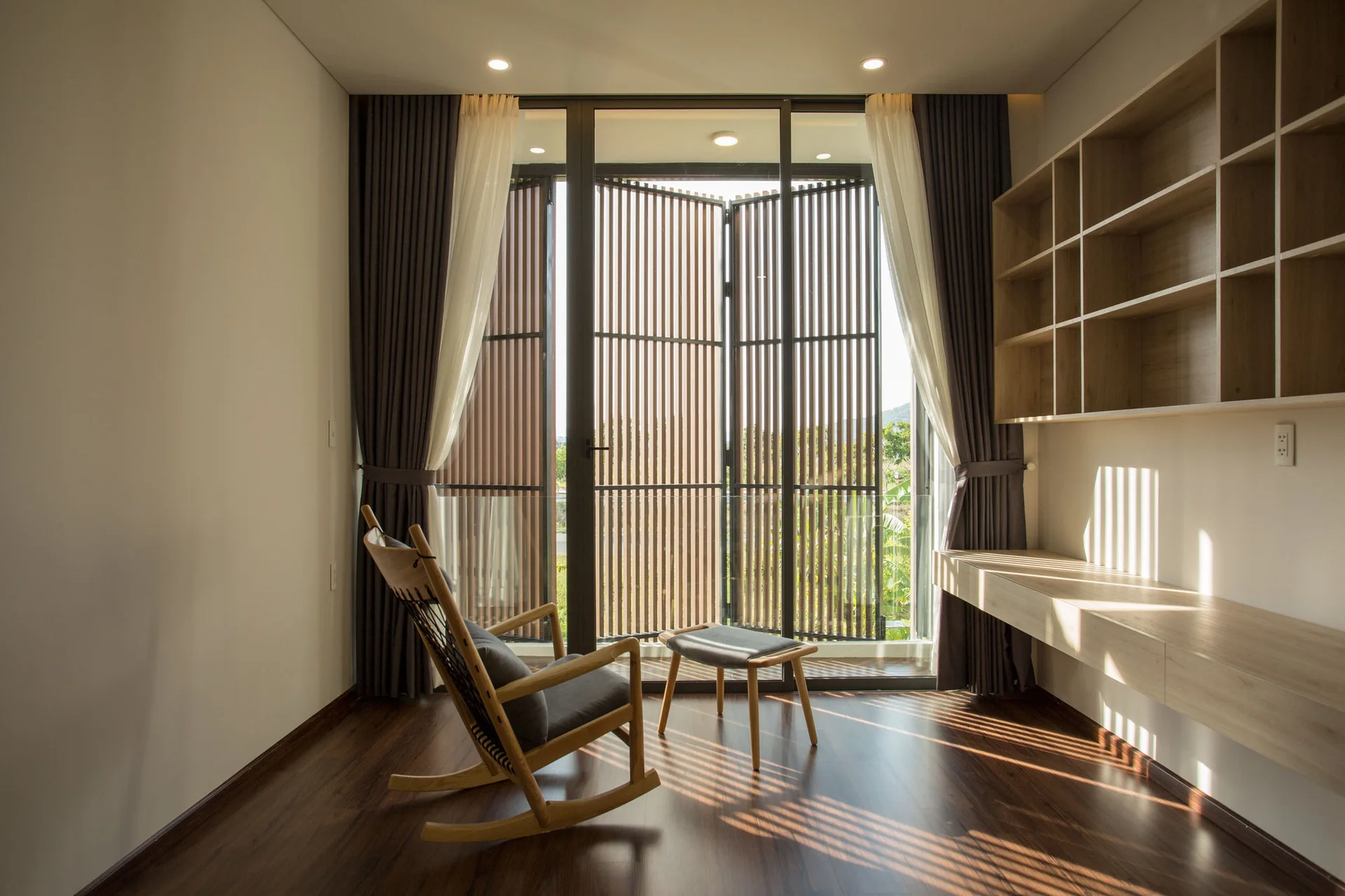 A wooden rocking chair and stool sit near a window with vertical wood slat screens, next to a floating desk and wall shelves on dark floors.