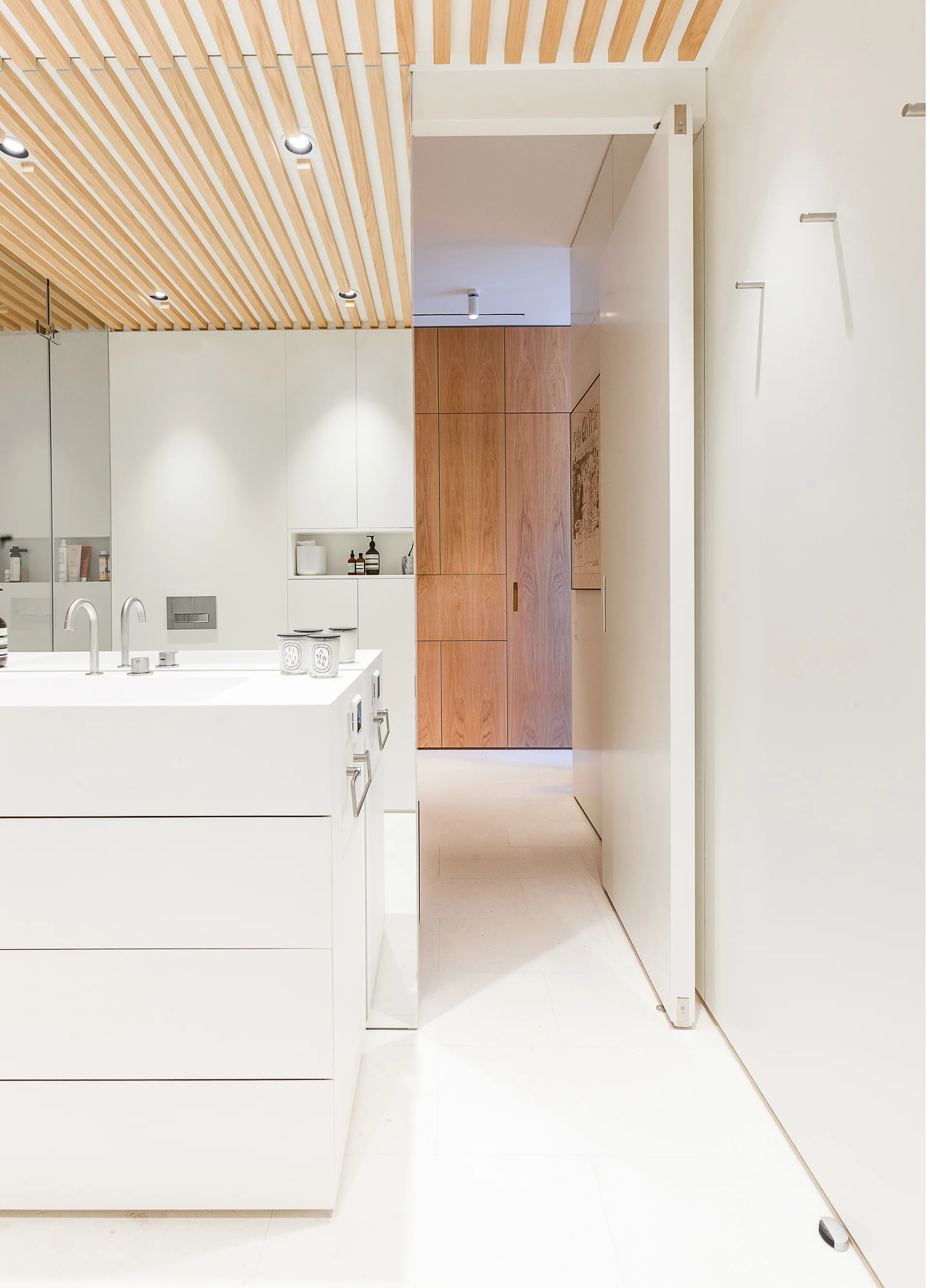 A minimalist white bathroom featuring a modern vanity, chrome faucets, a wood-slatted ceiling with recessed lighting, and a tall pivot door.