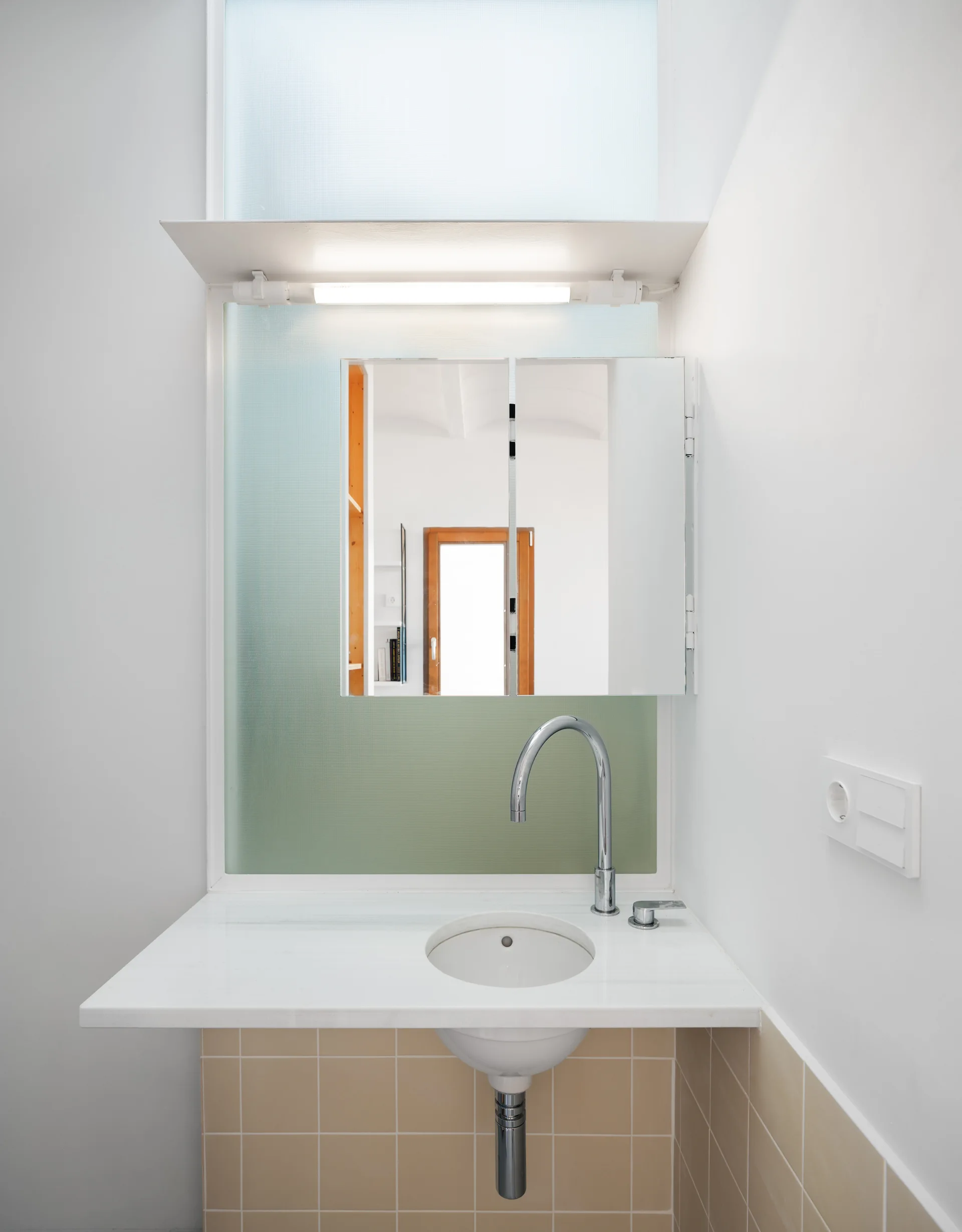 A wall-mounted white vanity with a circular integrated sink set against beige square tiles under a mirrored cabinet and frosted glass pane.