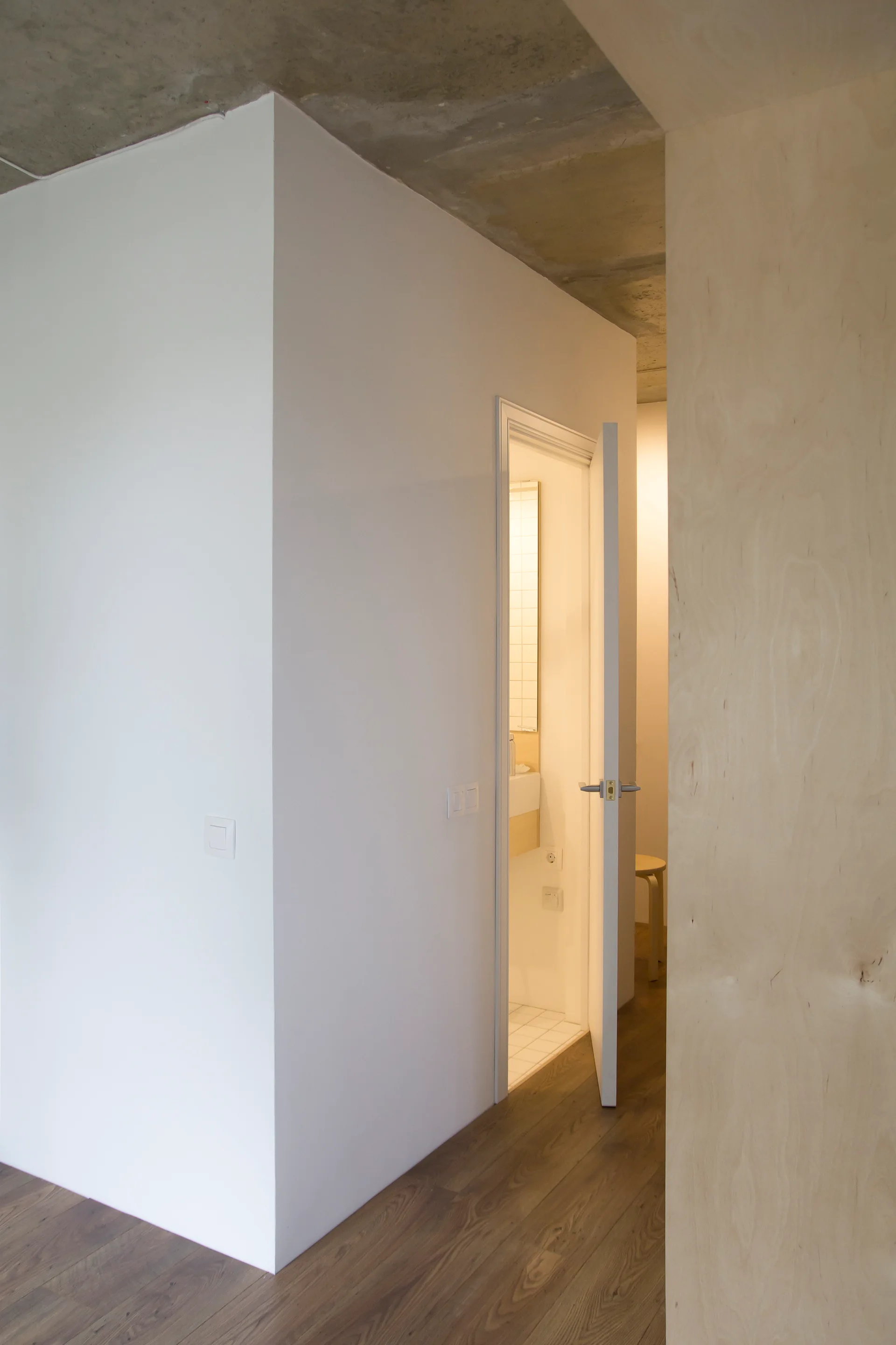 A minimalist hallway features a white partition wall, an exposed concrete ceiling, wood floors, and a plywood paneled wall.