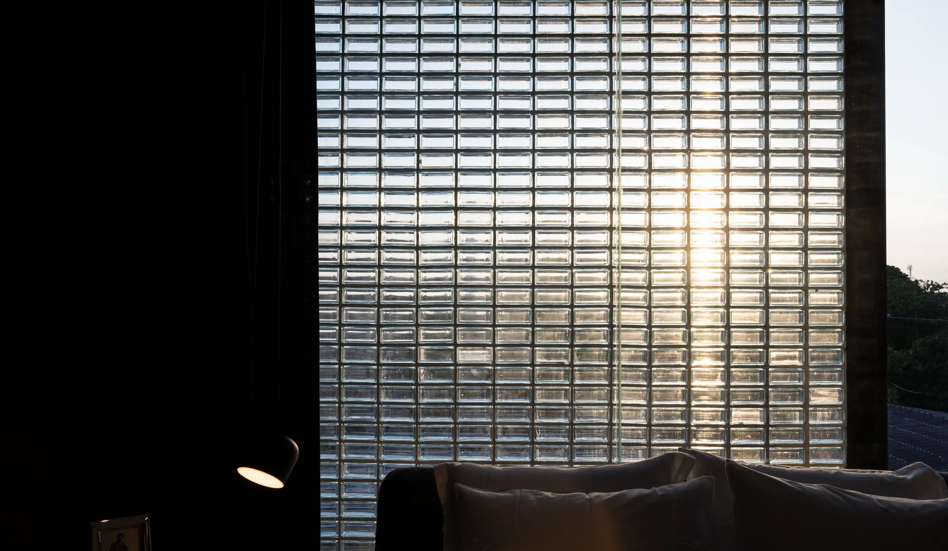Sunlight filters through a large glass block wall in a dark modern bedroom with white pillows in the foreground.
