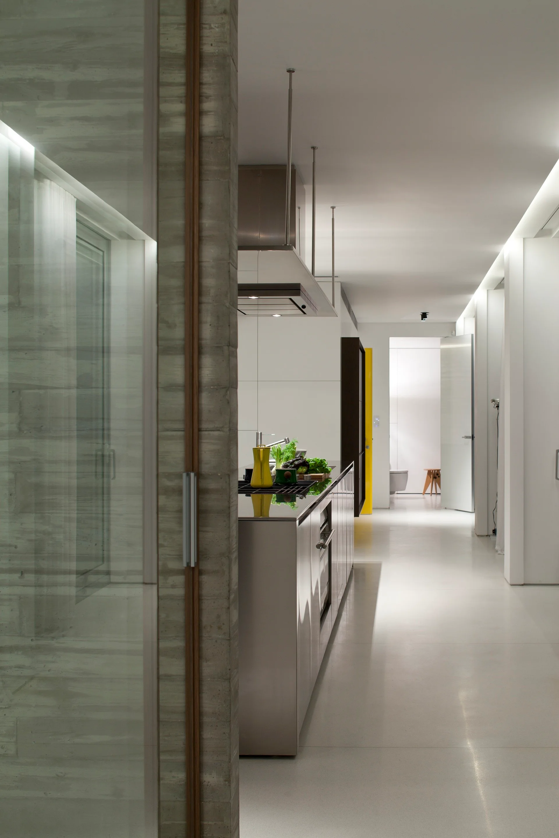 A modern minimalist kitchen corridor showcasing a sleek stainless steel island, white cabinetry, and a concrete partition wall.