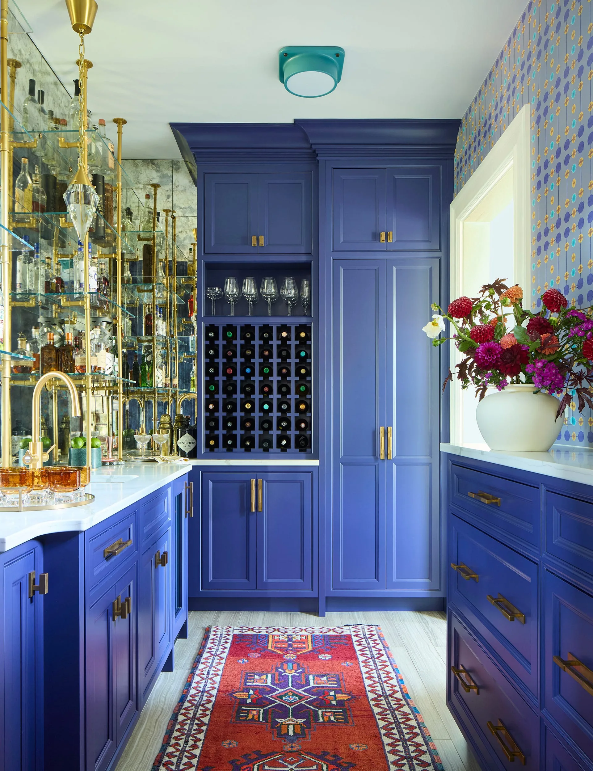 A royal blue home bar features matching cabinetry, an integrated wine rack, glass shelves with brass supports, and a patterned runner rug.