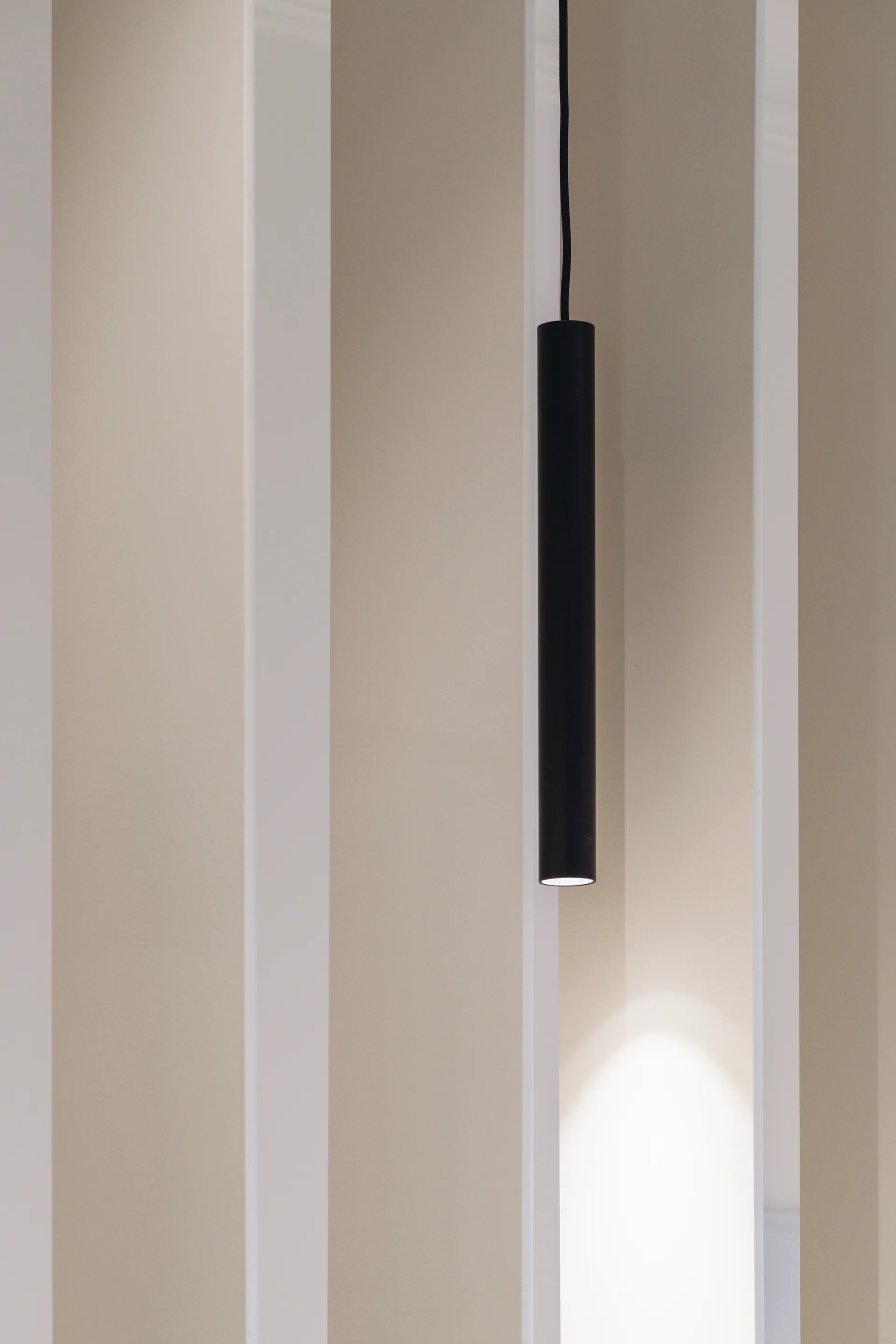A sleek matte black cylinder pendant light hangs vertically against a neutral beige wall with prominent vertical slats, casting a warm downlight.