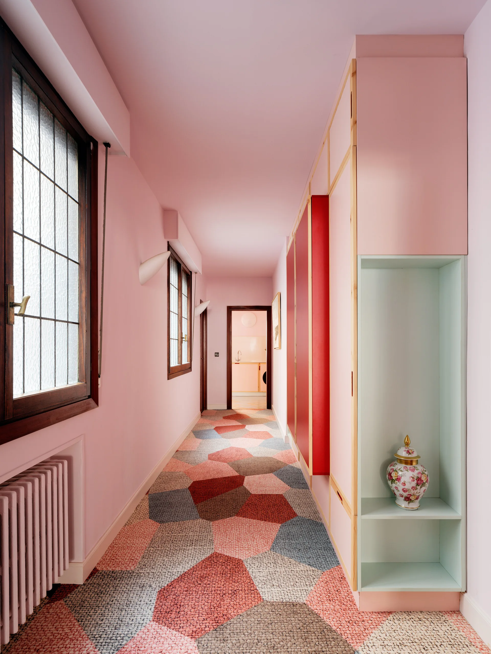 A long residential hallway featuring pink walls and ceiling, custom cabinetry with red doors, and a bold hexagonal pattern carpet.