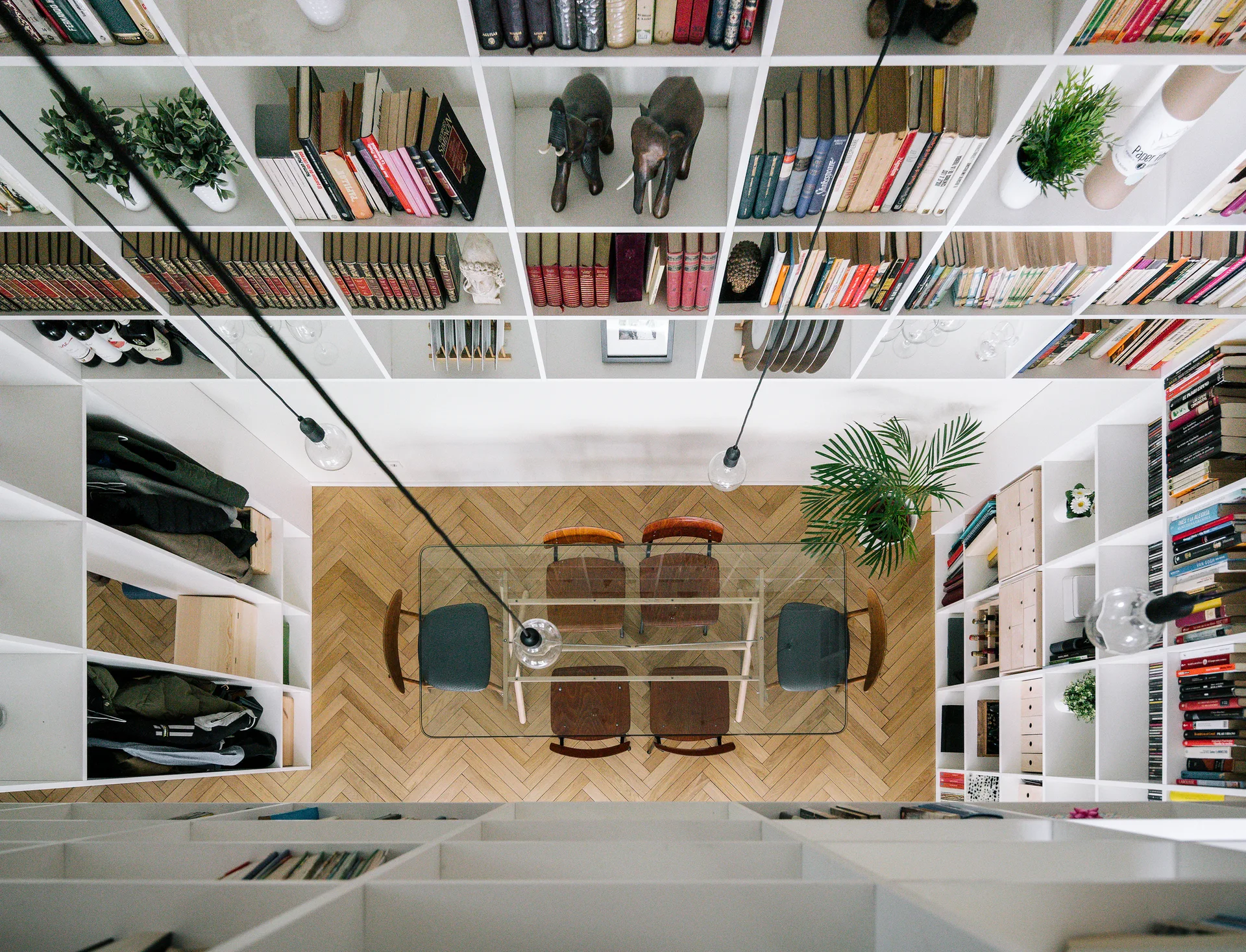 An overhead view of a glass dining table and chairs set on herringbone wood flooring, surrounded by white grid-patterned built-in bookcases.