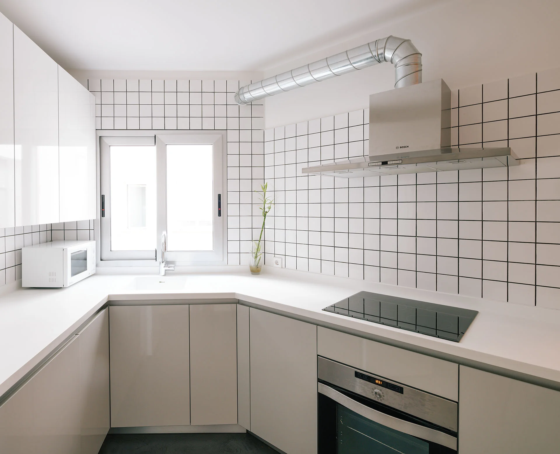 A minimalist L-shaped kitchen featuring white and grey cabinets, a square grid tile backsplash, and a stainless steel range hood with exposed ductwork.