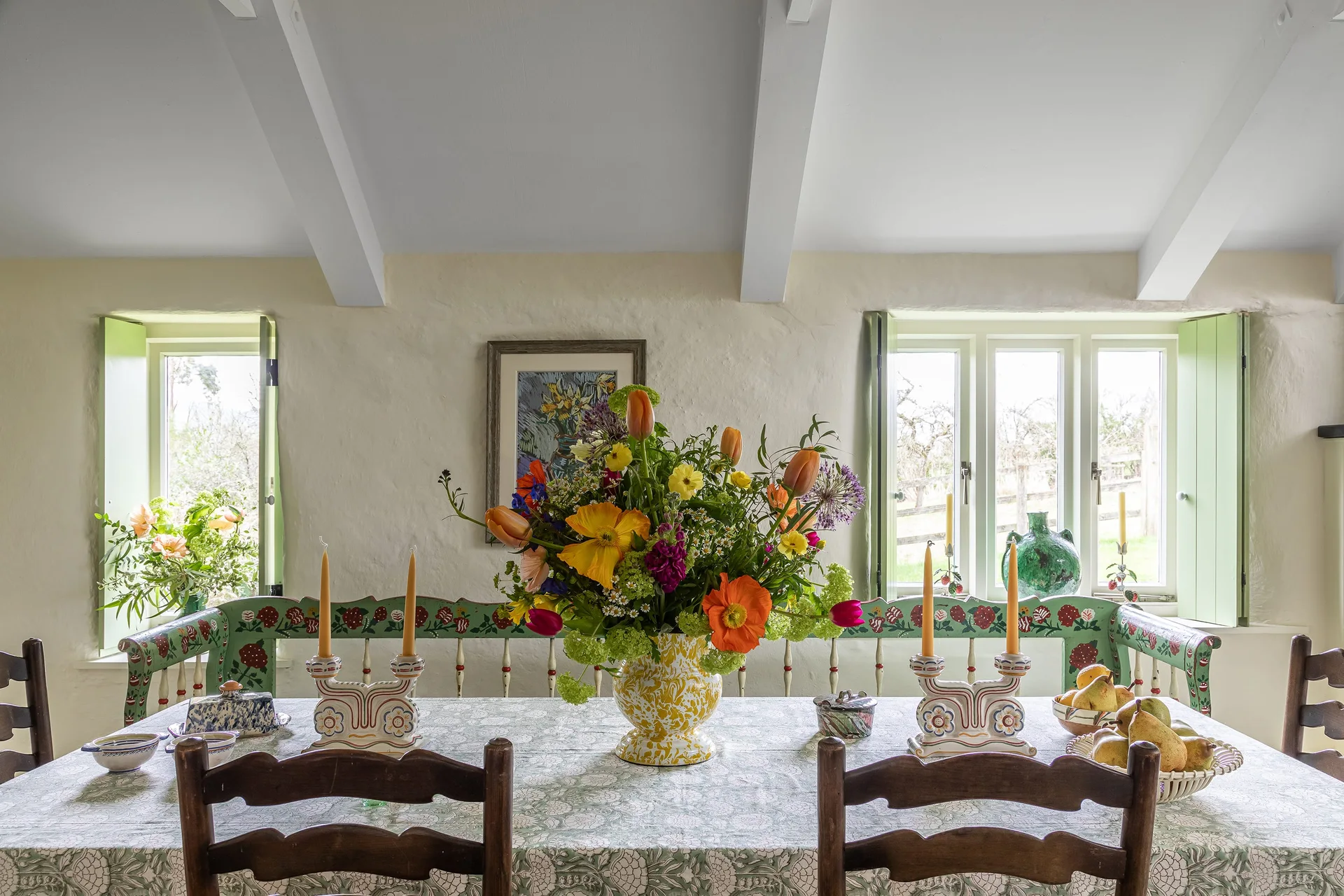 A rustic dining table with a botanical tablecloth and wild flowers sits in front of a hand-painted green bench and shuttered windows.