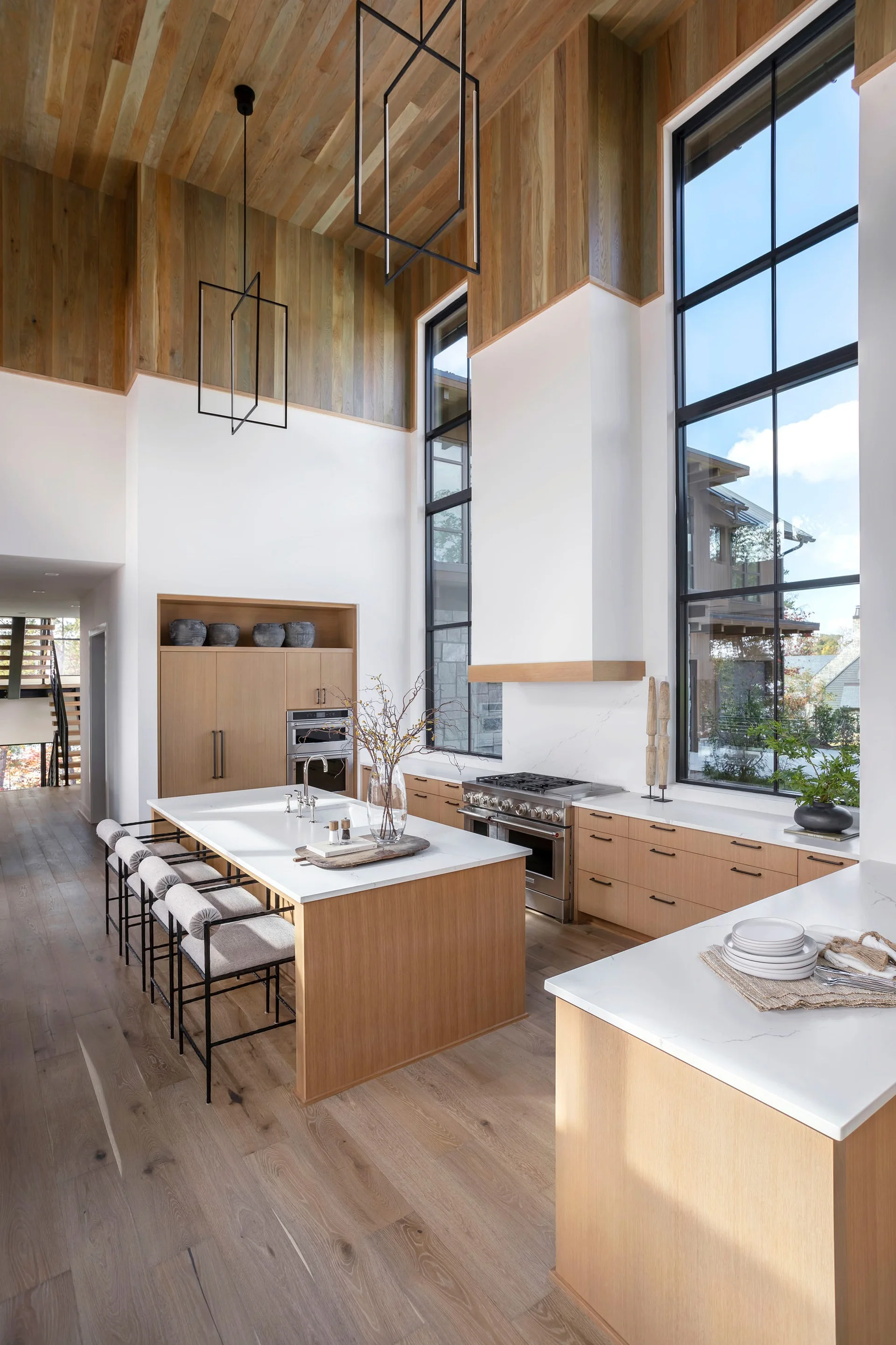 A bright high-ceiling kitchen featuring a light wood island with modern stools, white countertops, tall steel-framed windows, and a wood-paneled ceiling.
